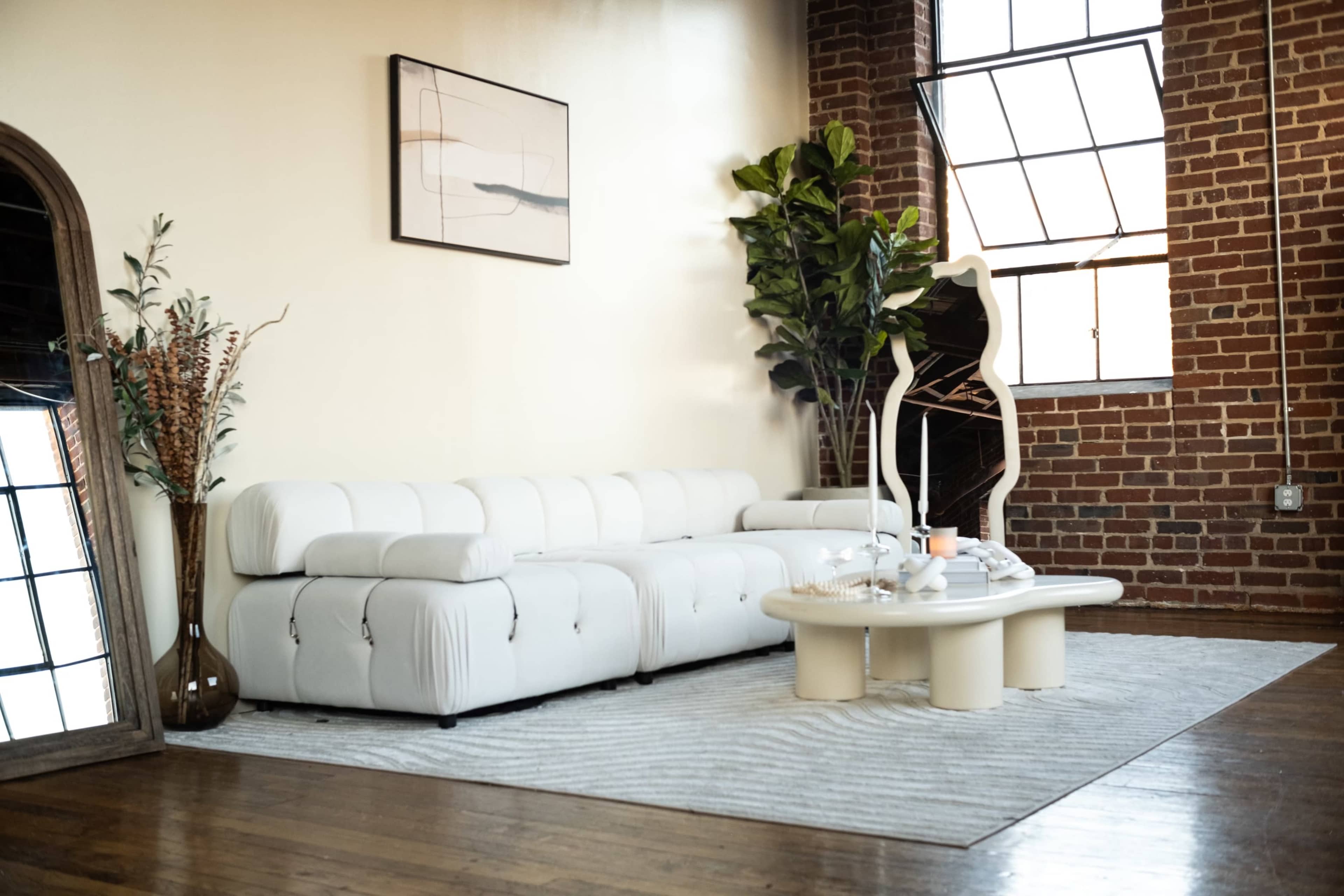 A modern living room features a white sectional sofa, a round coffee table, and a large window with exposed brick walls in the background.