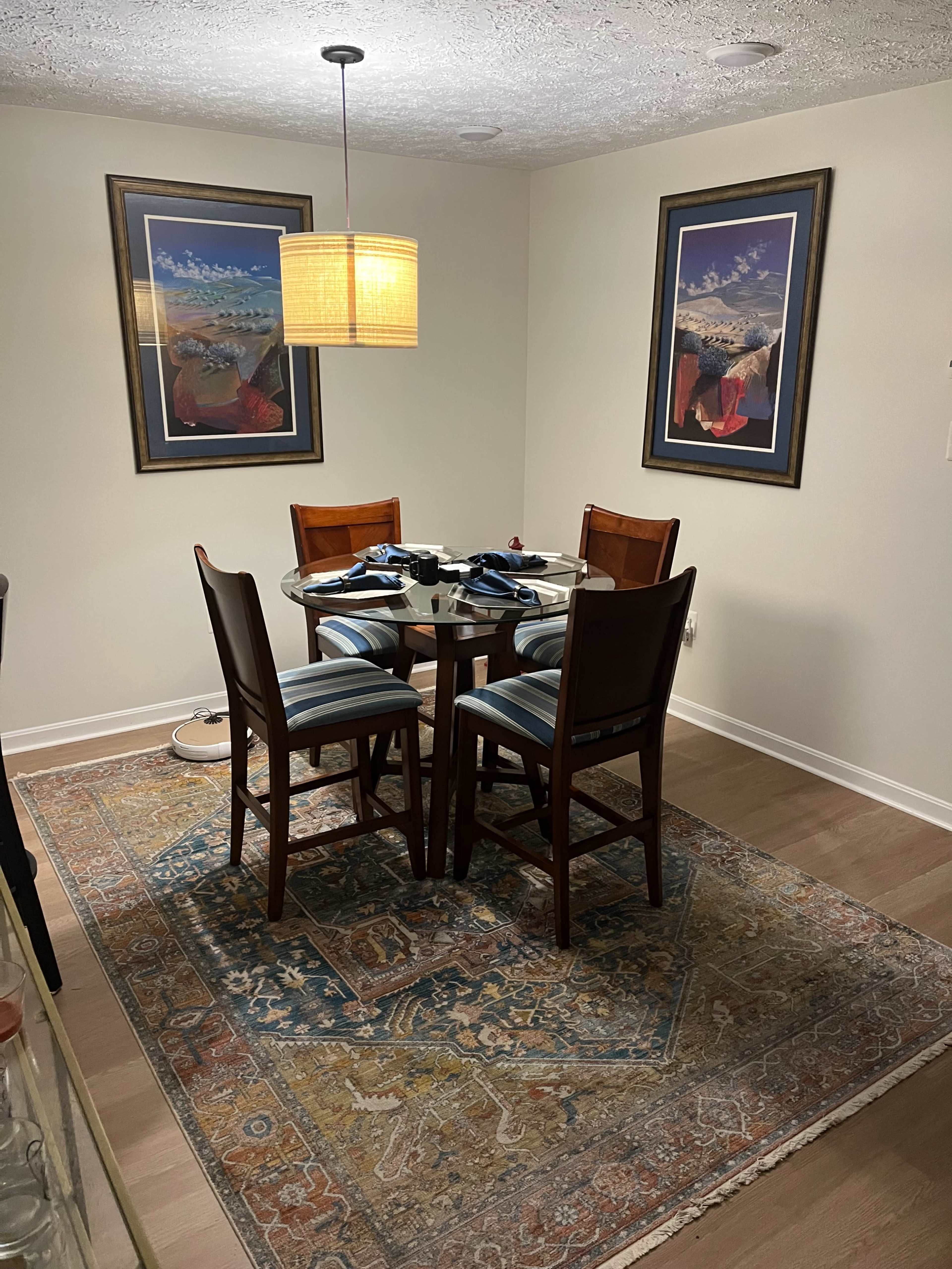 A dining area with a round glass table surrounded by four wooden chairs, set on a patterned rug, and illuminated by a pendant light.