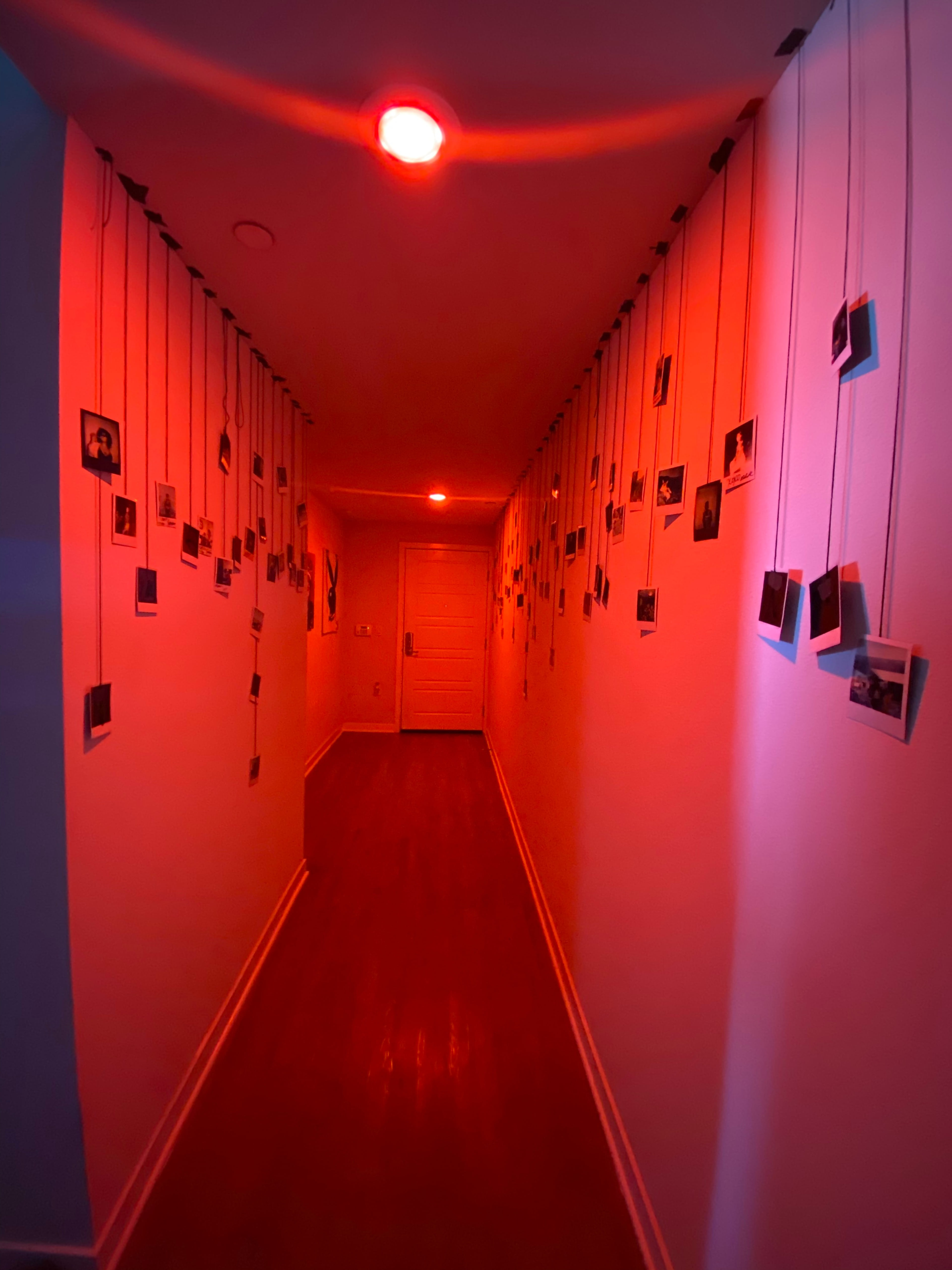 A narrow hallway is illuminated by a red light, with framed photographs hanging from strings along the walls.