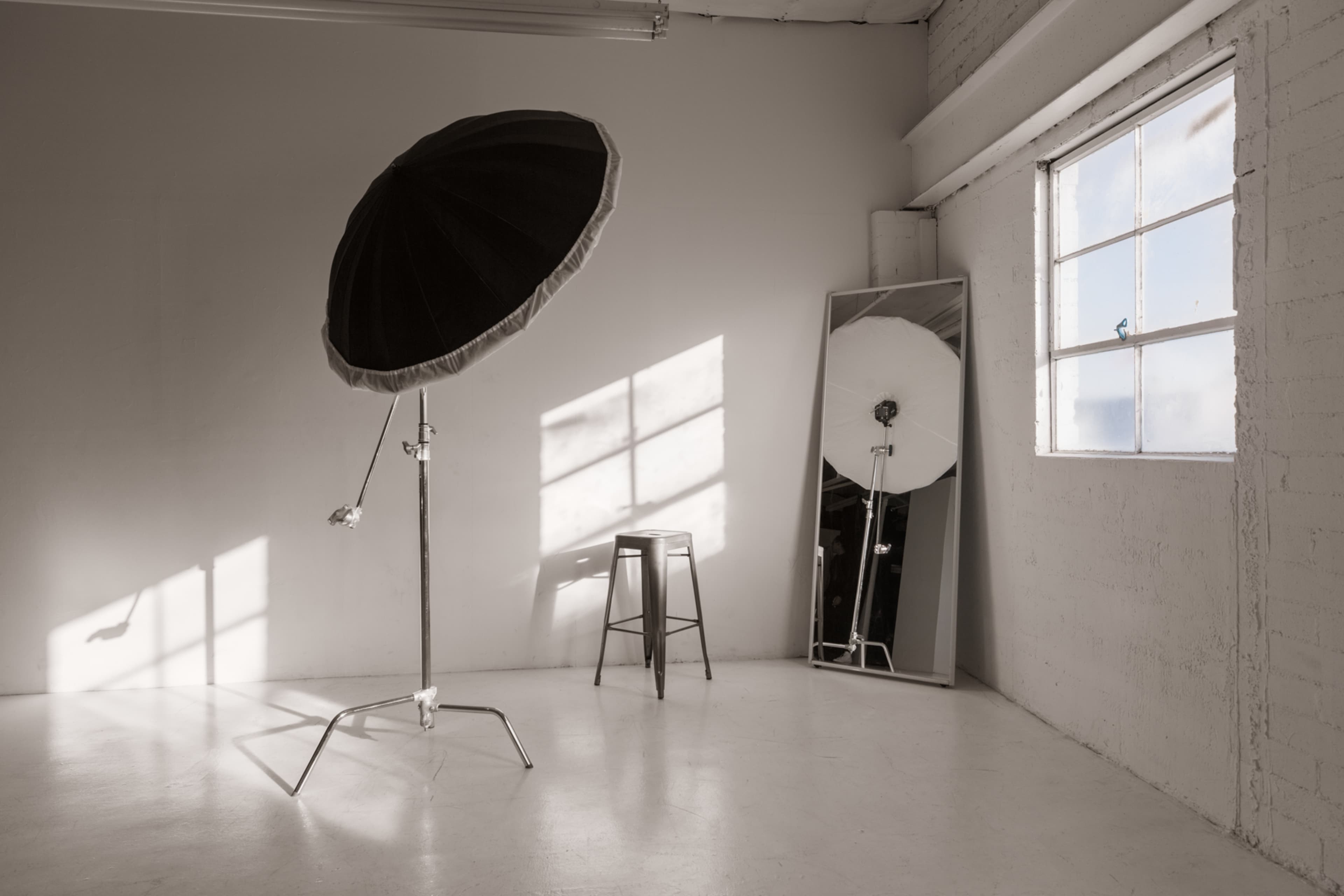 A photography studio features a large softbox, a mirrored wall, a stool, and sunlight casting shadows on the floor.