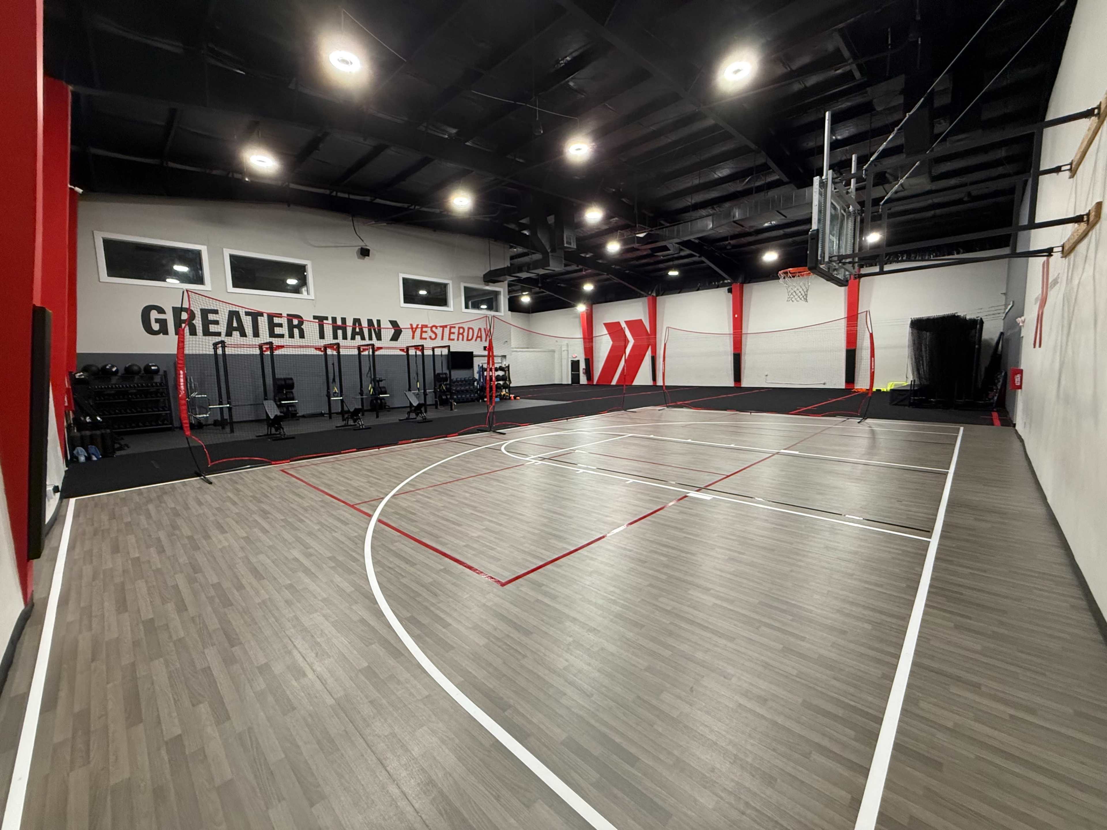 The image shows an indoor basketball court with a wooden floor, red and white markings, and fitness equipment along the walls.