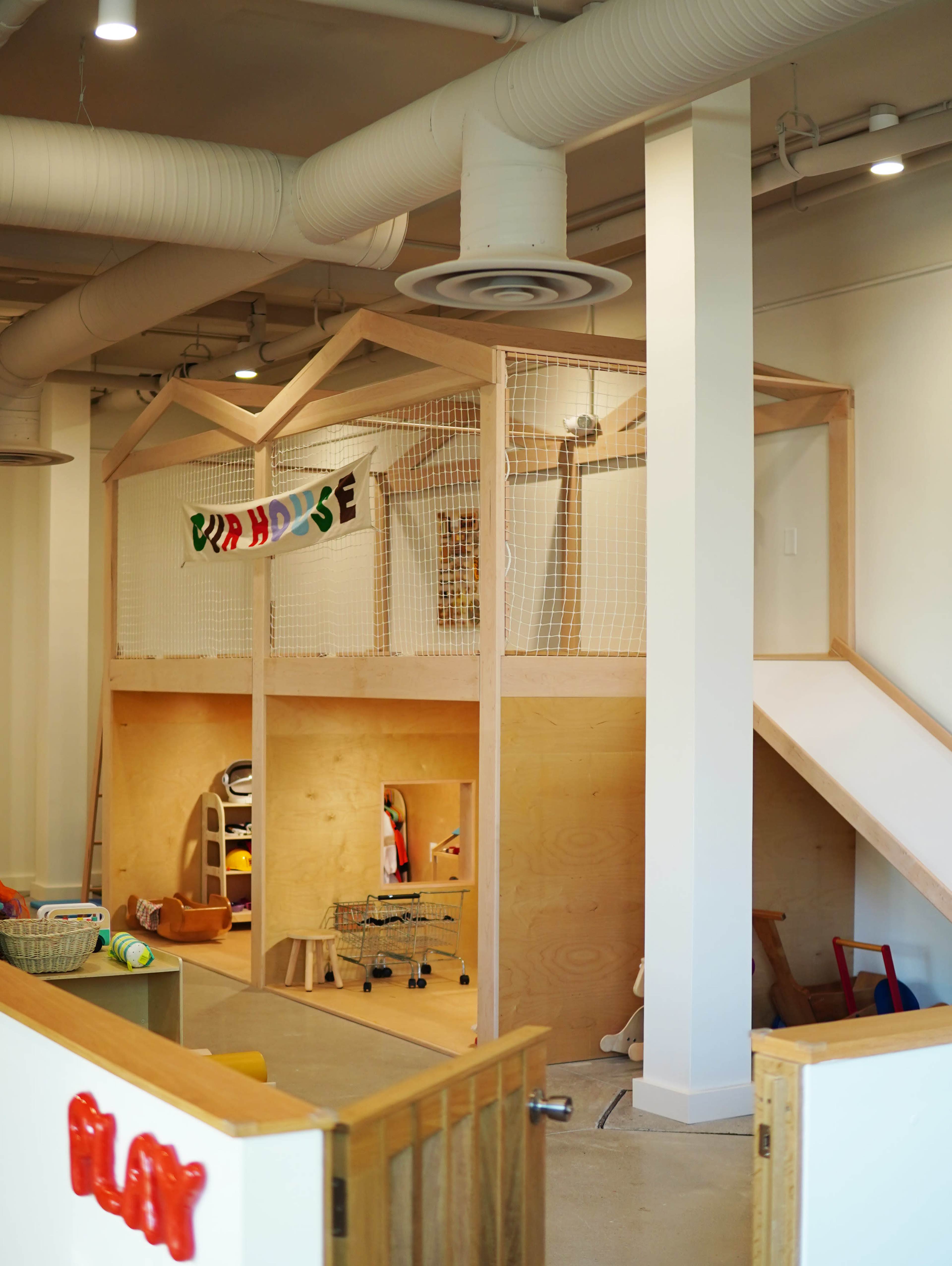 The image shows a spacious indoor play area featuring a wooden playhouse structure with a netted enclosure, adjacent to a staircase and various toys scattered around.