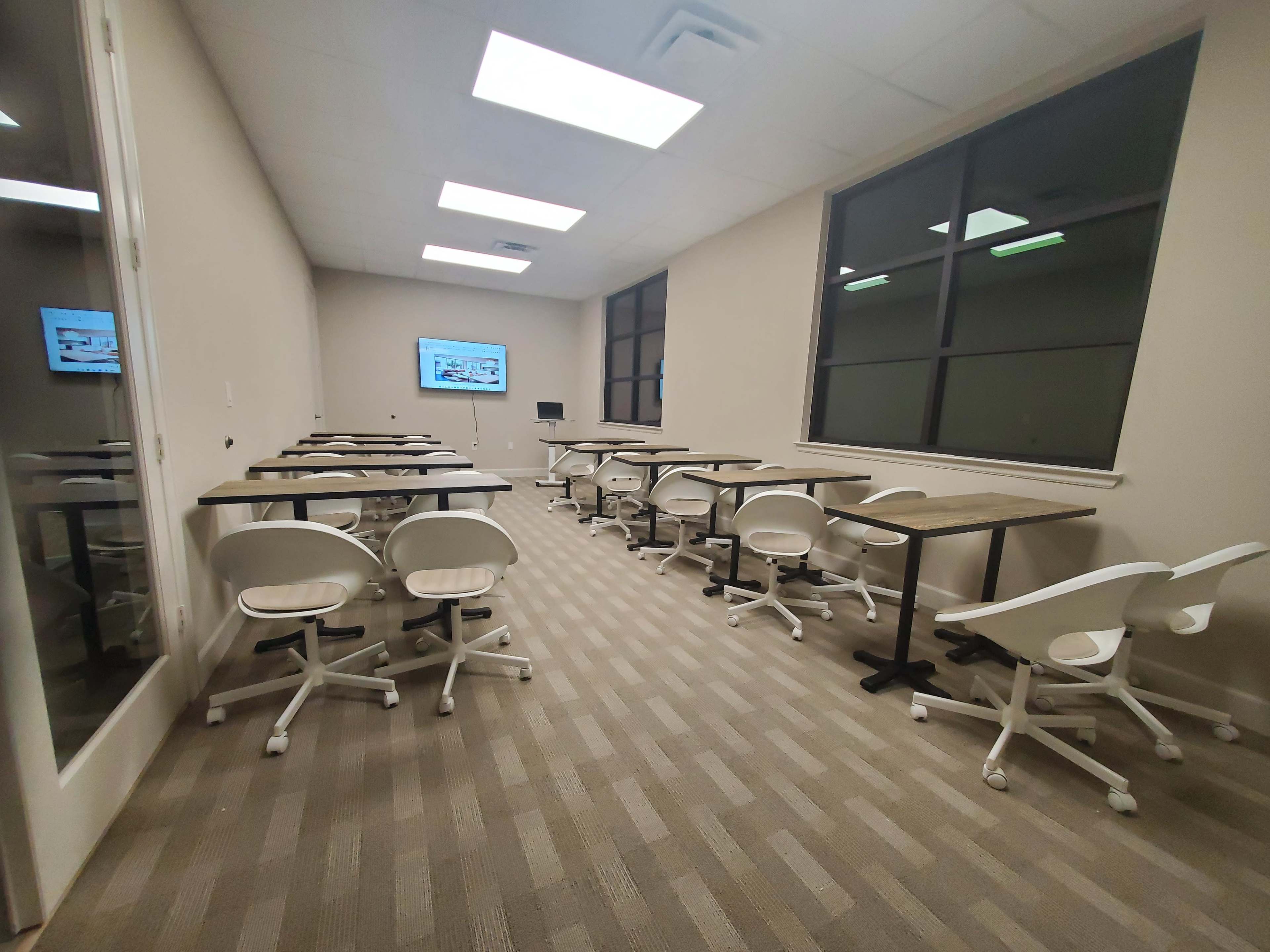 A classroom is set up with several desks and rolling chairs facing a large screen at the front.