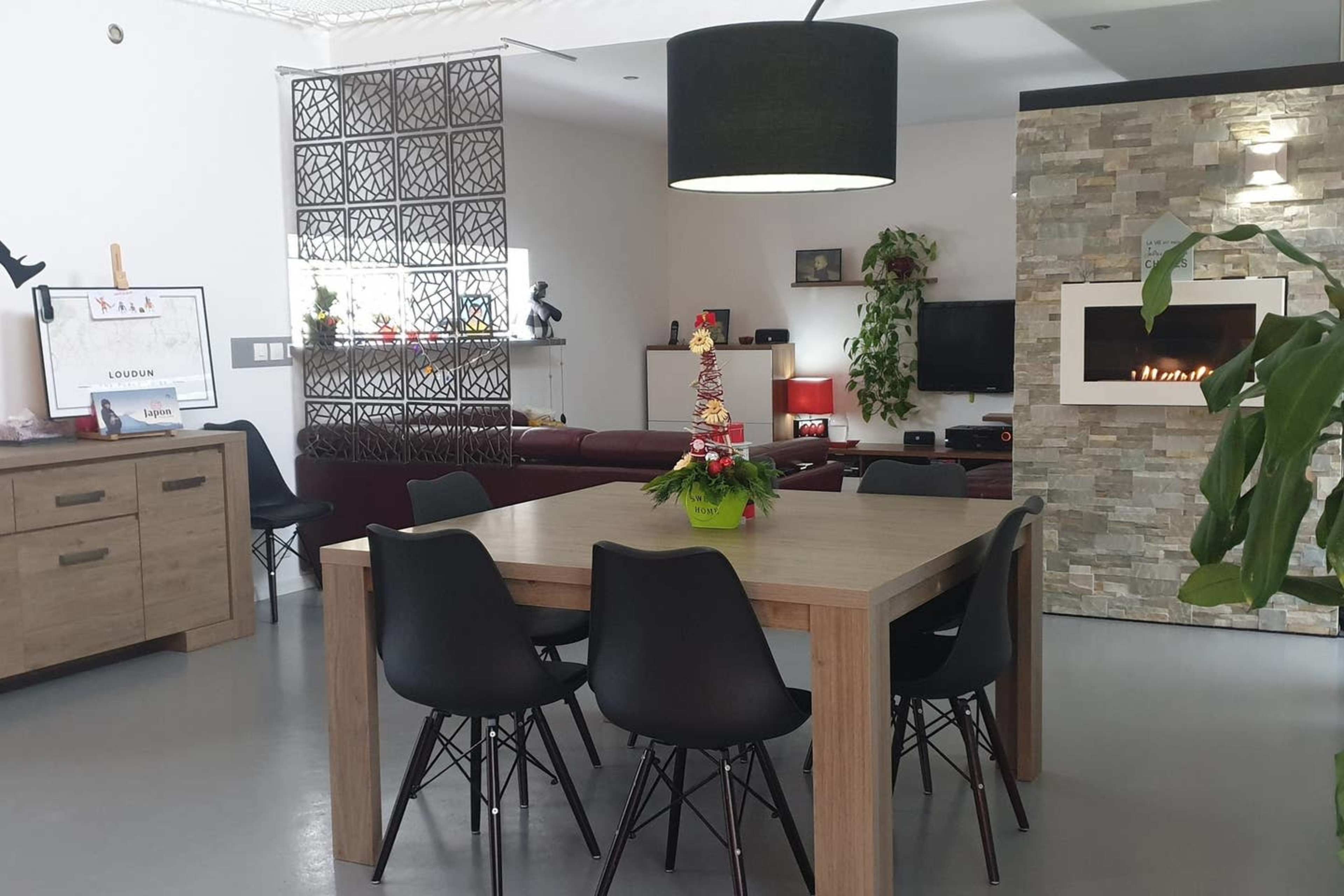 The image shows a modern dining area with a wooden table surrounded by black chairs, set against a backdrop of a living space featuring a stone fireplace and decorative plants.