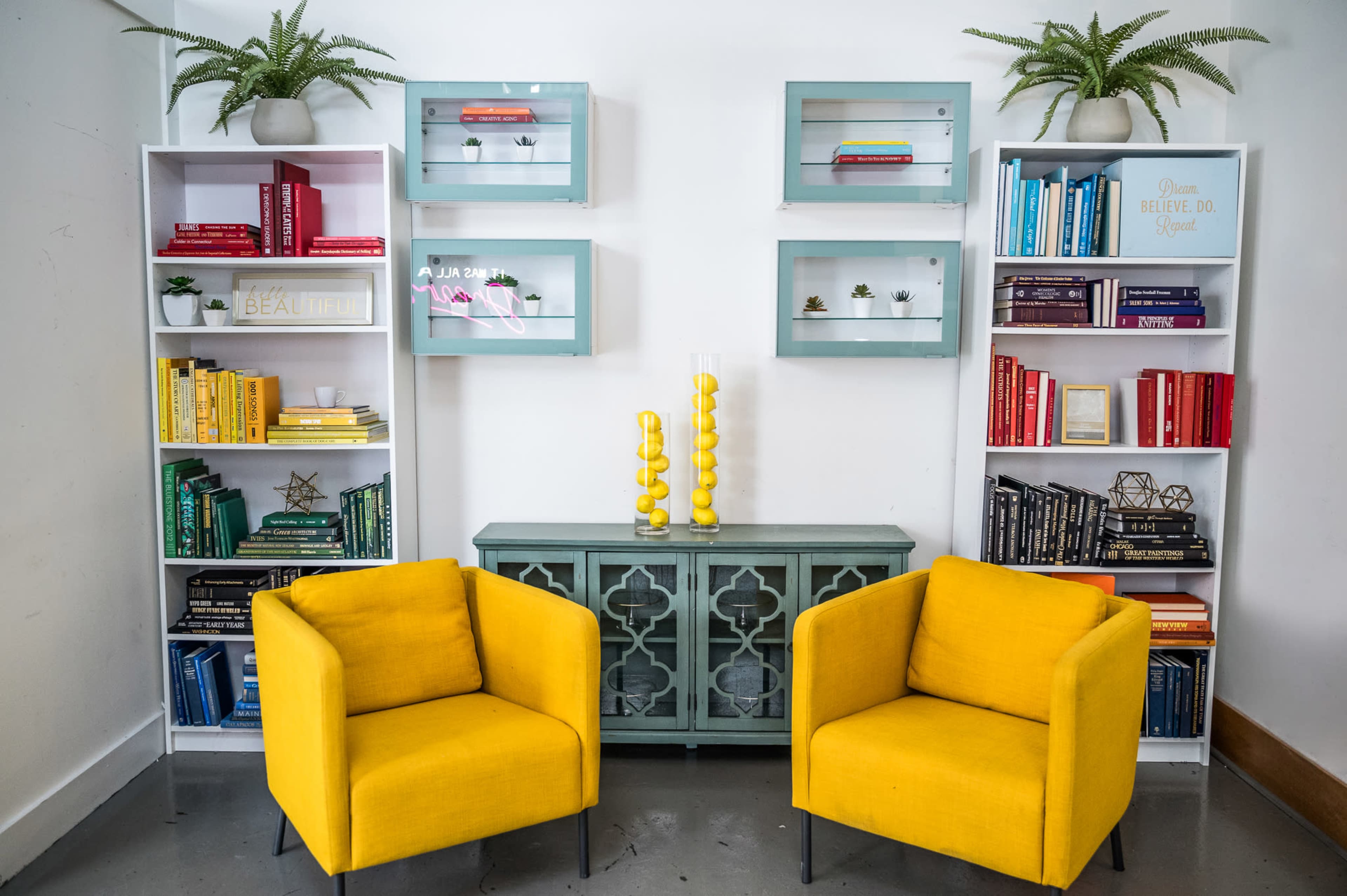 A vibrant seating area features two yellow chairs flanking a teal side table, surrounded by white bookshelves filled with colorful books and decorative items.