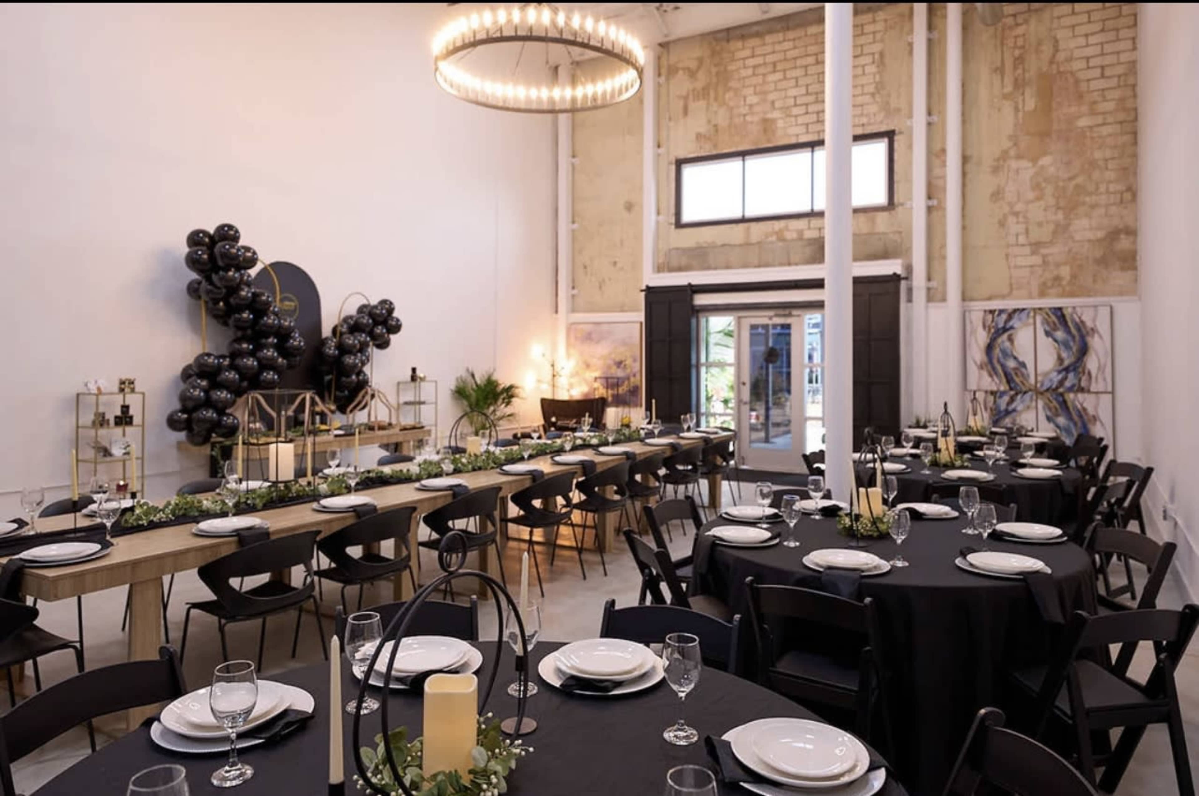 A spacious dining area set for an event, featuring long tables covered with black tablecloths and arranged with white plates, glassware, and decorative centerpieces.