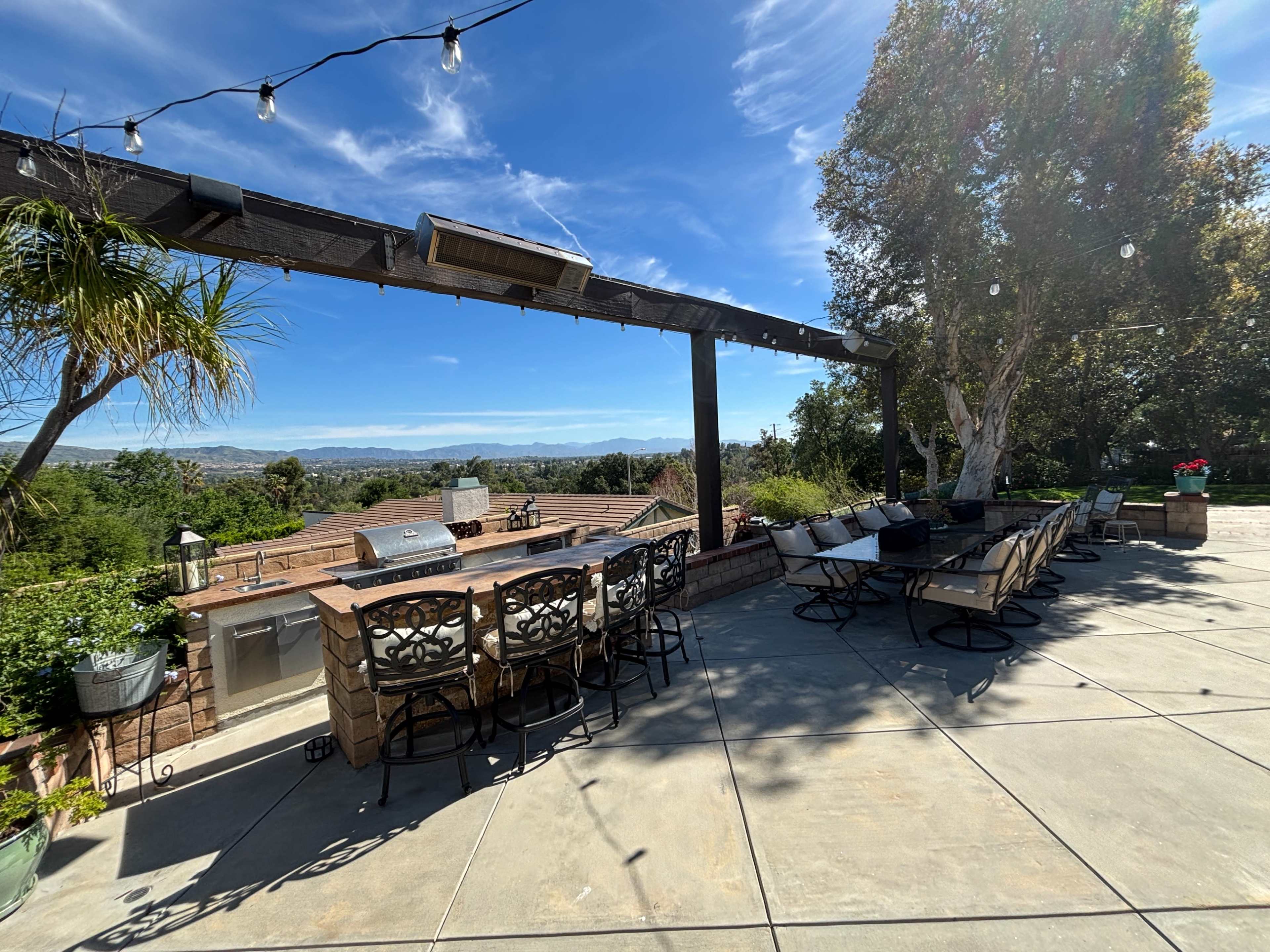 The image shows an outdoor patio area with bar seating and a grill, overlooking a scenic view of greenery and hills under a clear blue sky.