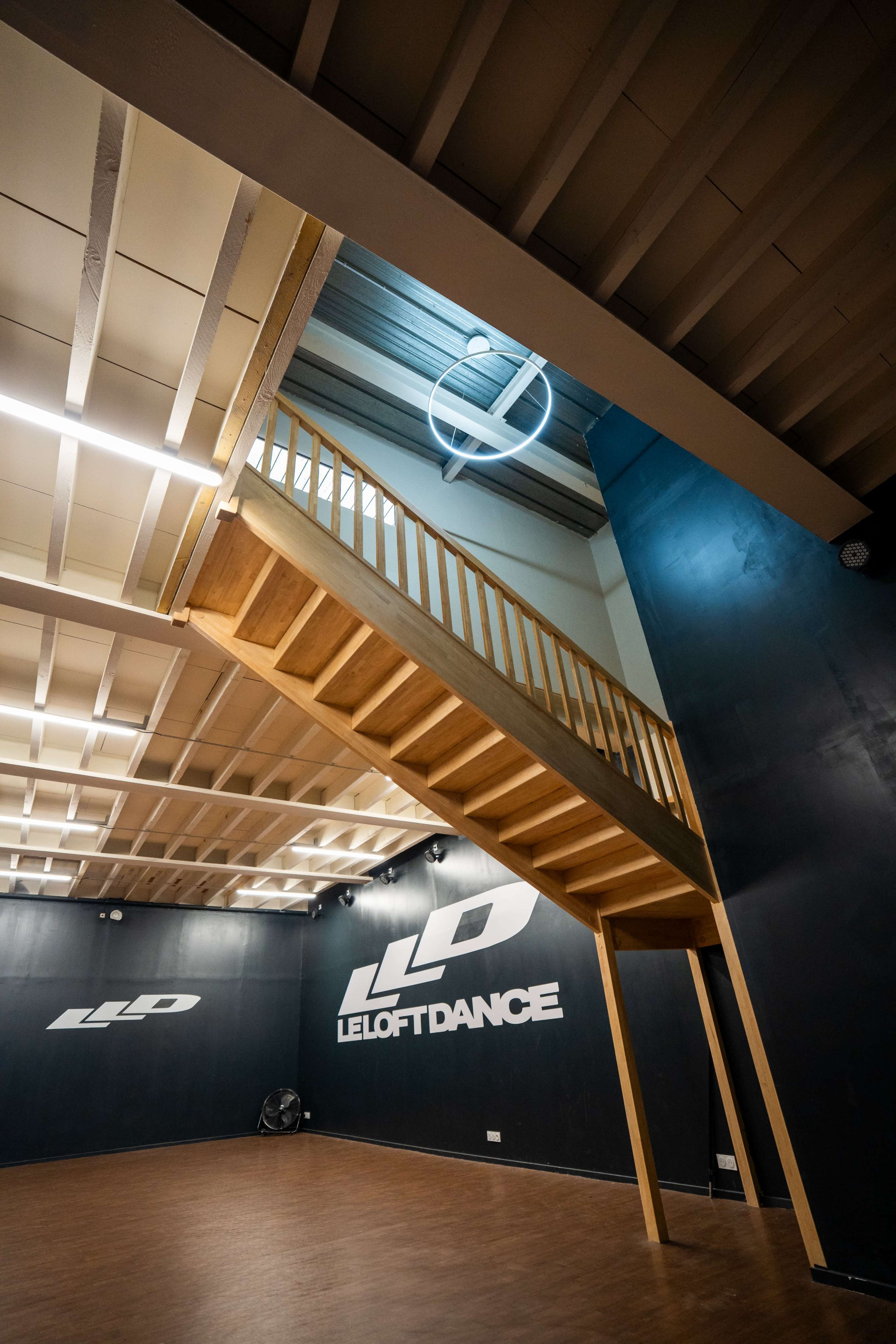 The image shows a dance studio with a prominent wooden staircase, dimly lit, and walls featuring the logo "LE LOFT DANCE."