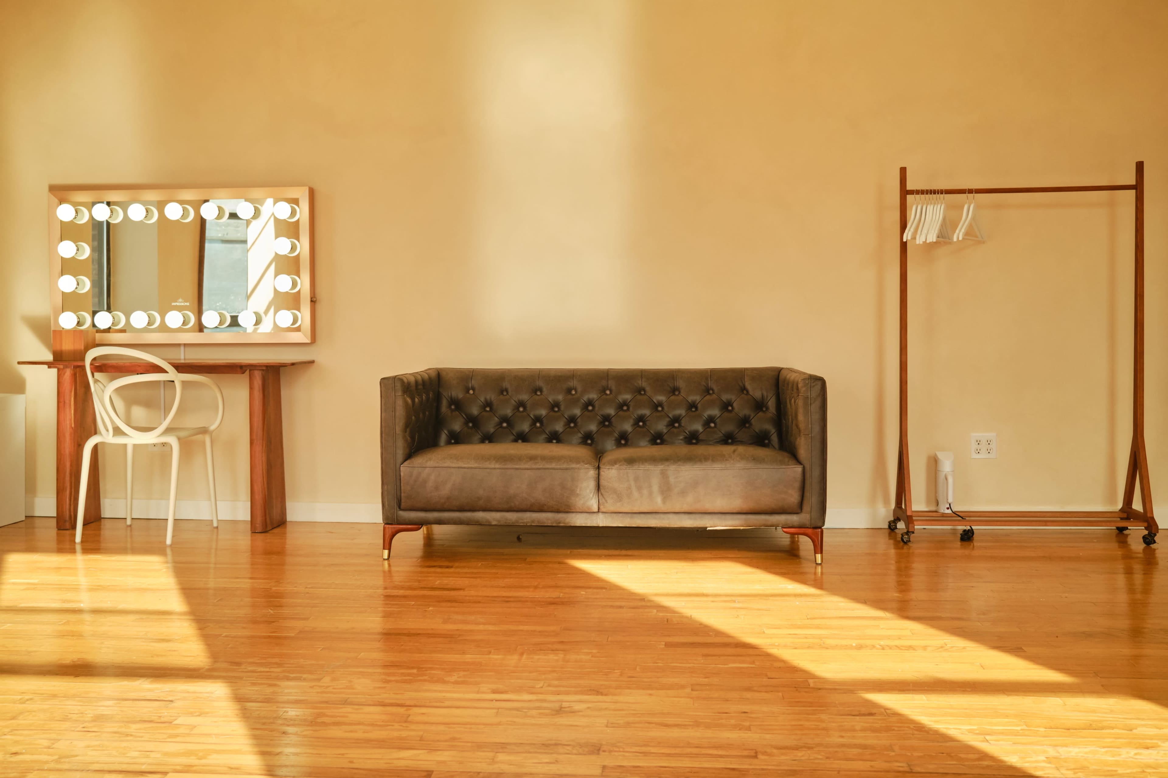 The scene features a cozy interior with a brown tufted sofa, a vanity mirror with lights, and a coat rack against a warm-toned wall, illuminated by natural light.