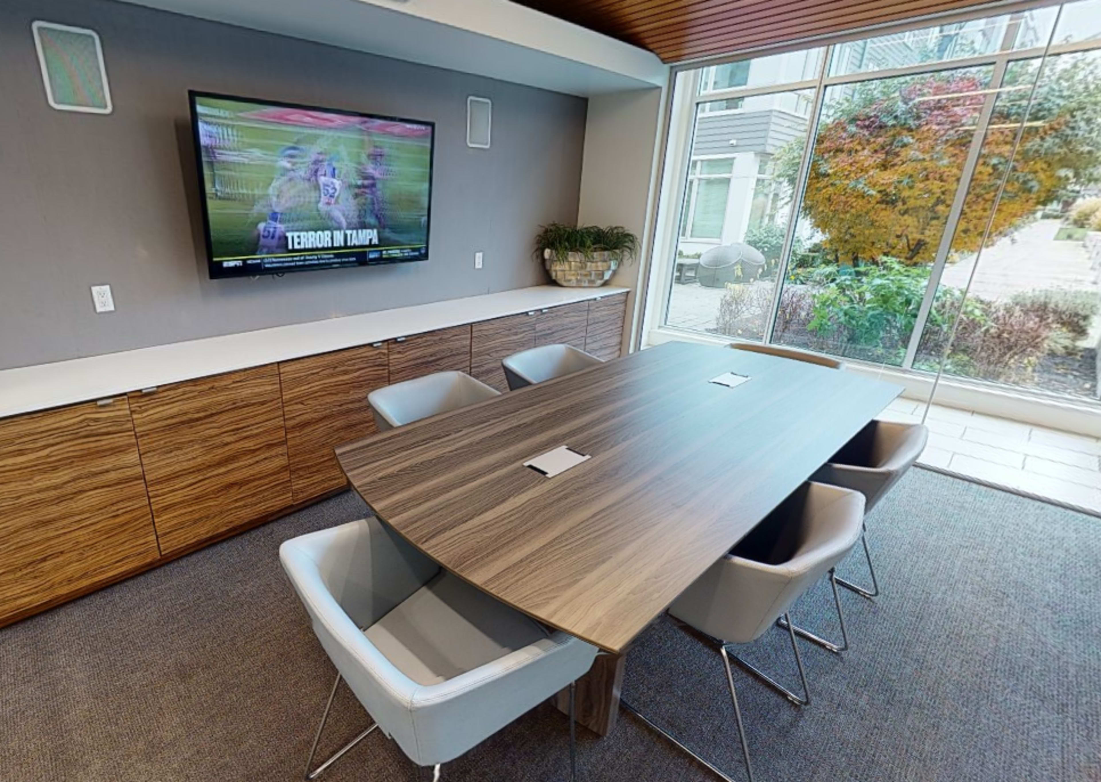 A conference room features a long wooden table surrounded by four chairs, with a television displaying a sports broadcast mounted on the wall.