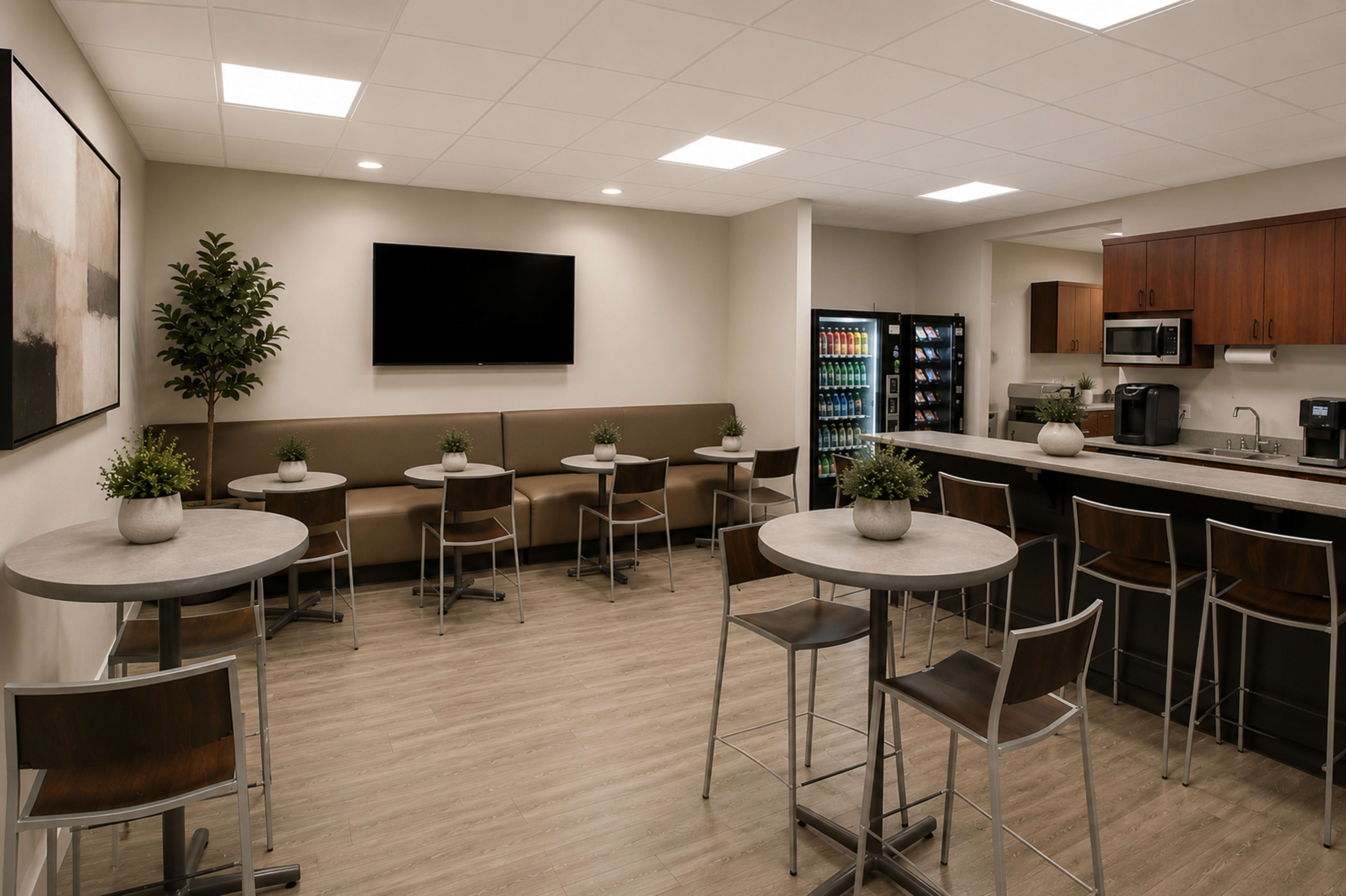 The image shows a modern break room with several small round tables, chairs, a kitchenette area, and a vending machine in the corner.