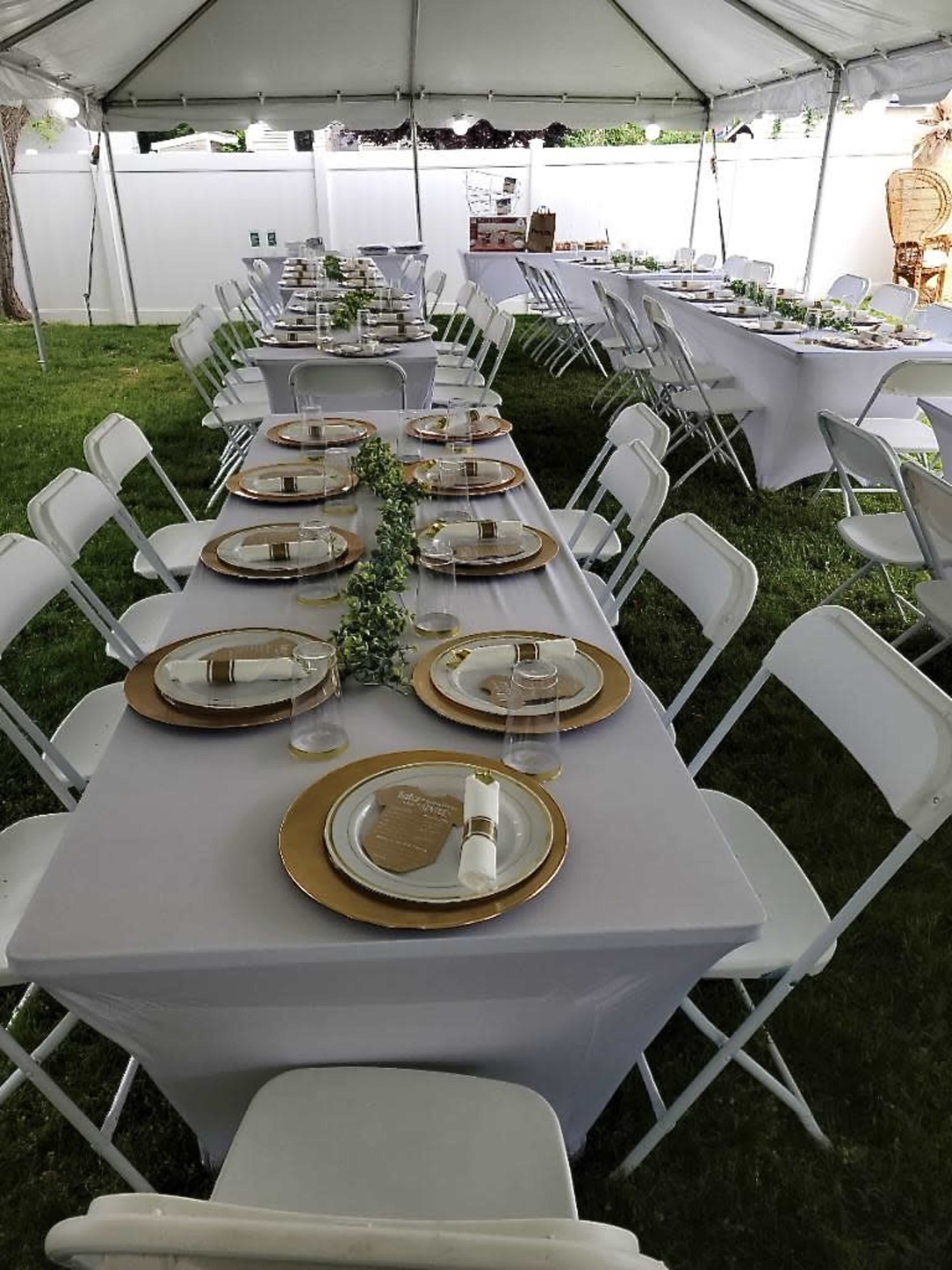 A tented outdoor space is set up with long tables covered in gray linens, featuring gold-rimmed plates and glassware arranged for a formal gathering.