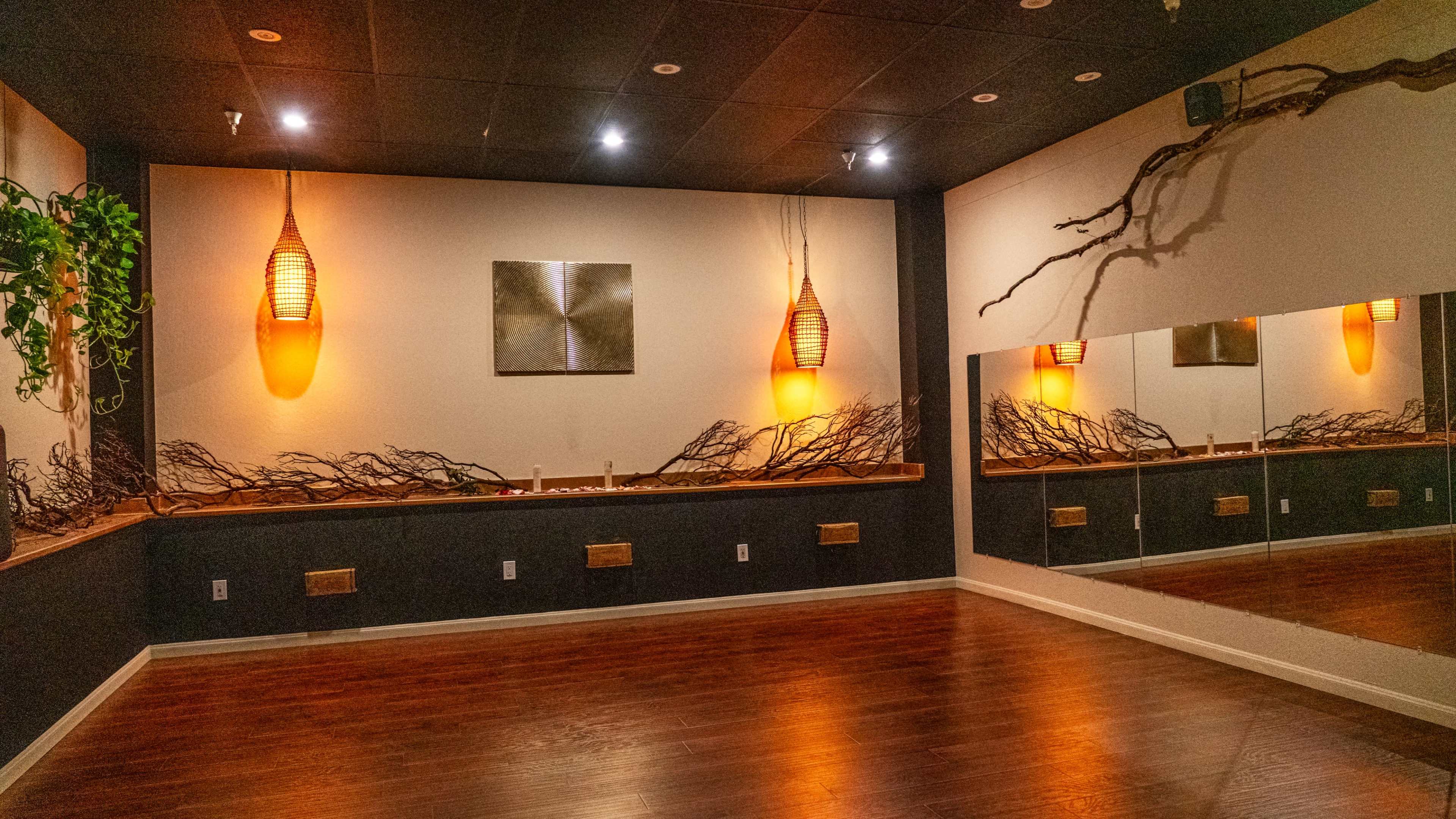 The image shows a spacious yoga studio with wooden floors, soft lighting from pendant lamps, and a mirrored wall featuring natural elements like branches.