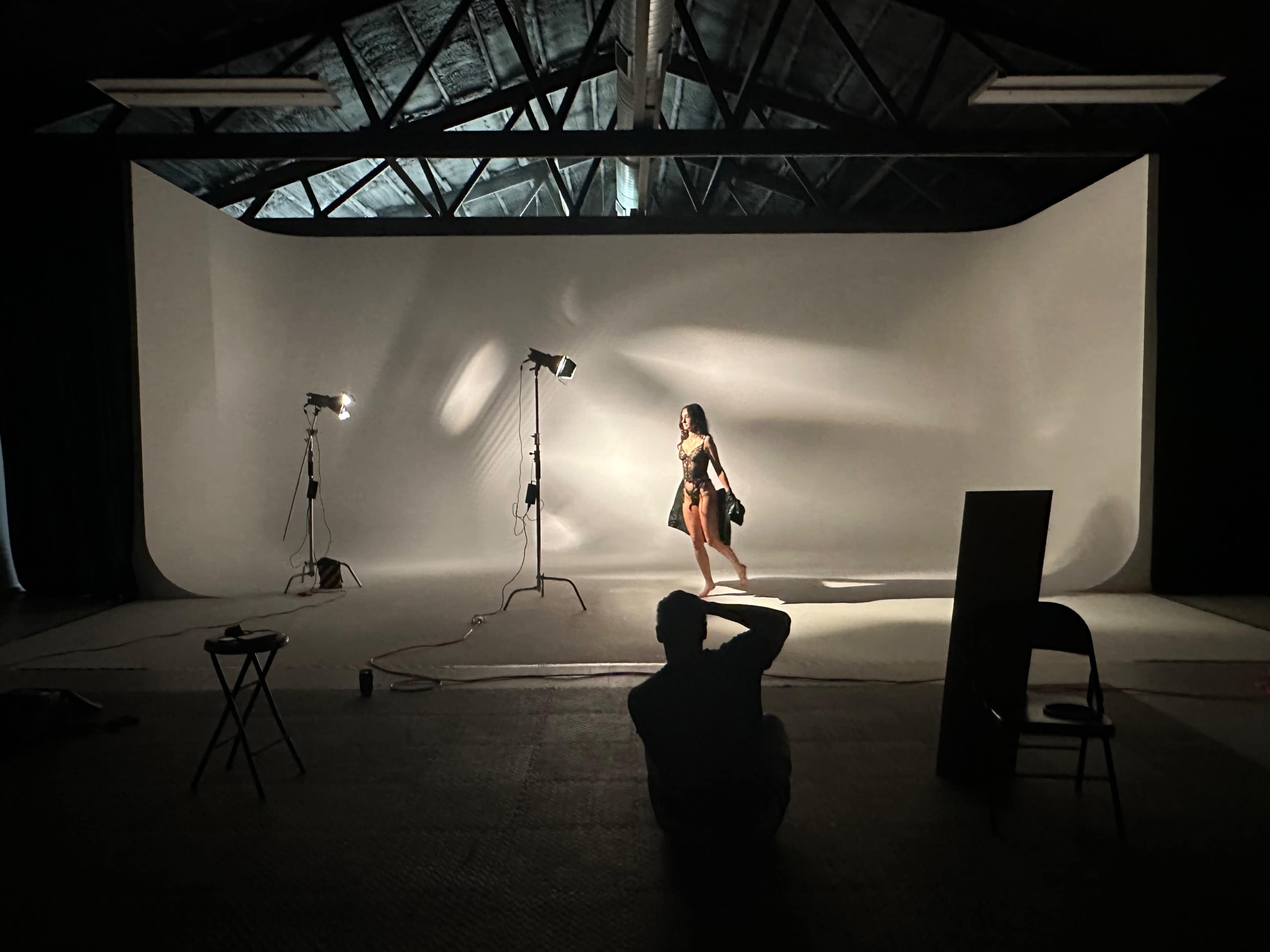 A person poses in a studio with a gray backdrop while a photographer captures the shot from a distance.