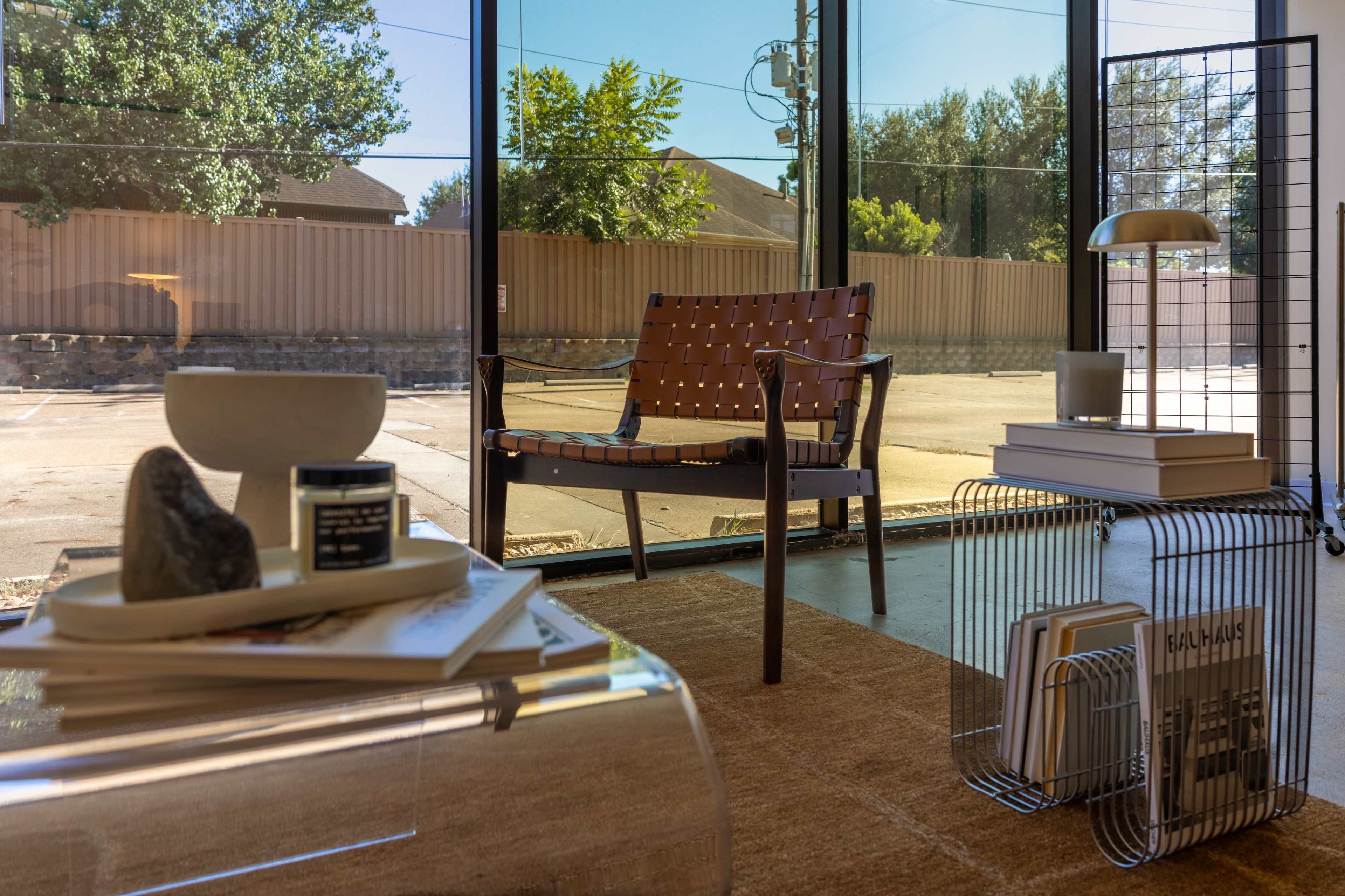 The image shows a modern interior with a brown leather chair, a glass coffee table with decor items, and large windows overlooking an outdoor area.