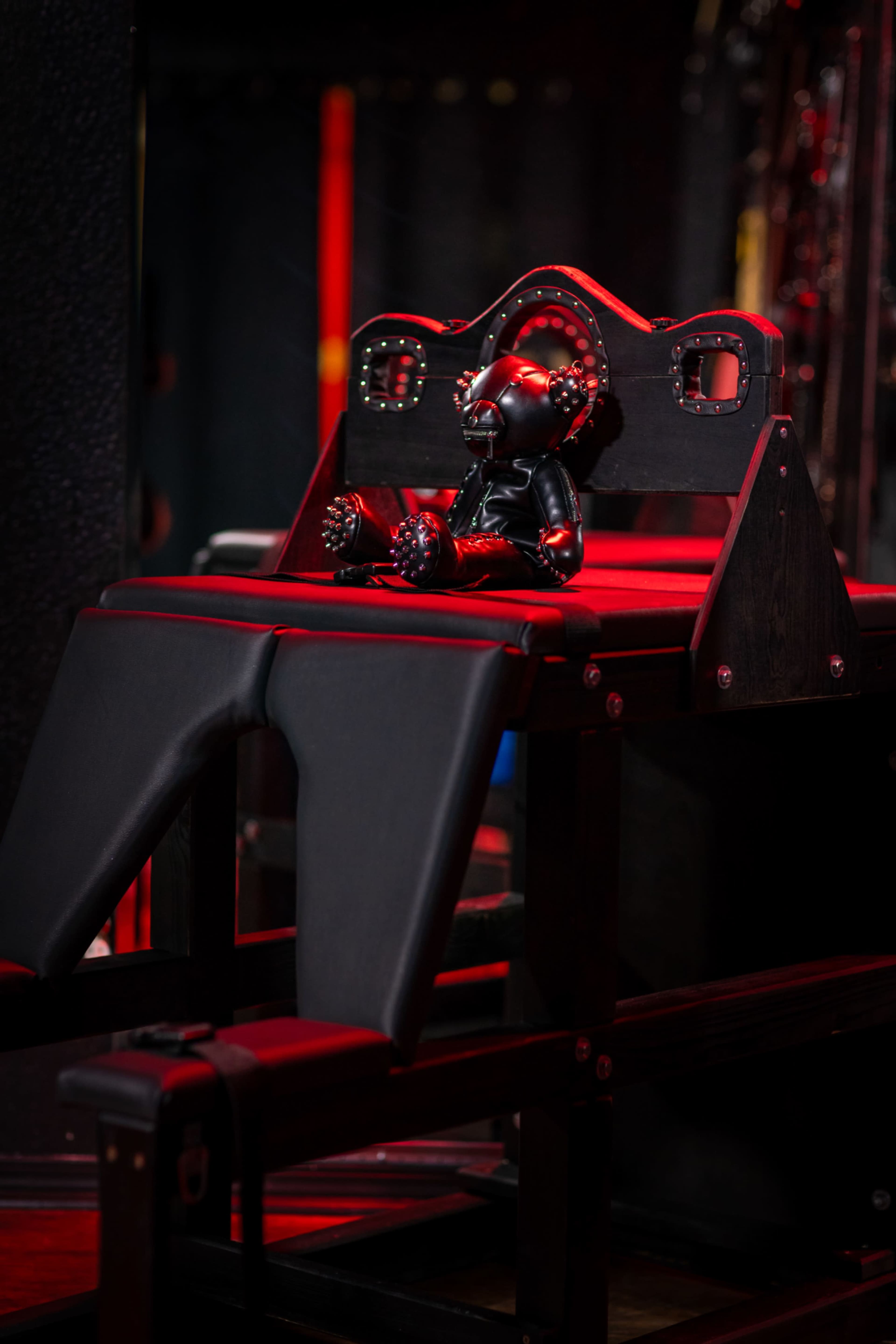 The image features a padded bondage bench with a small, plush doll seated on it, surrounded by dim red lighting.
