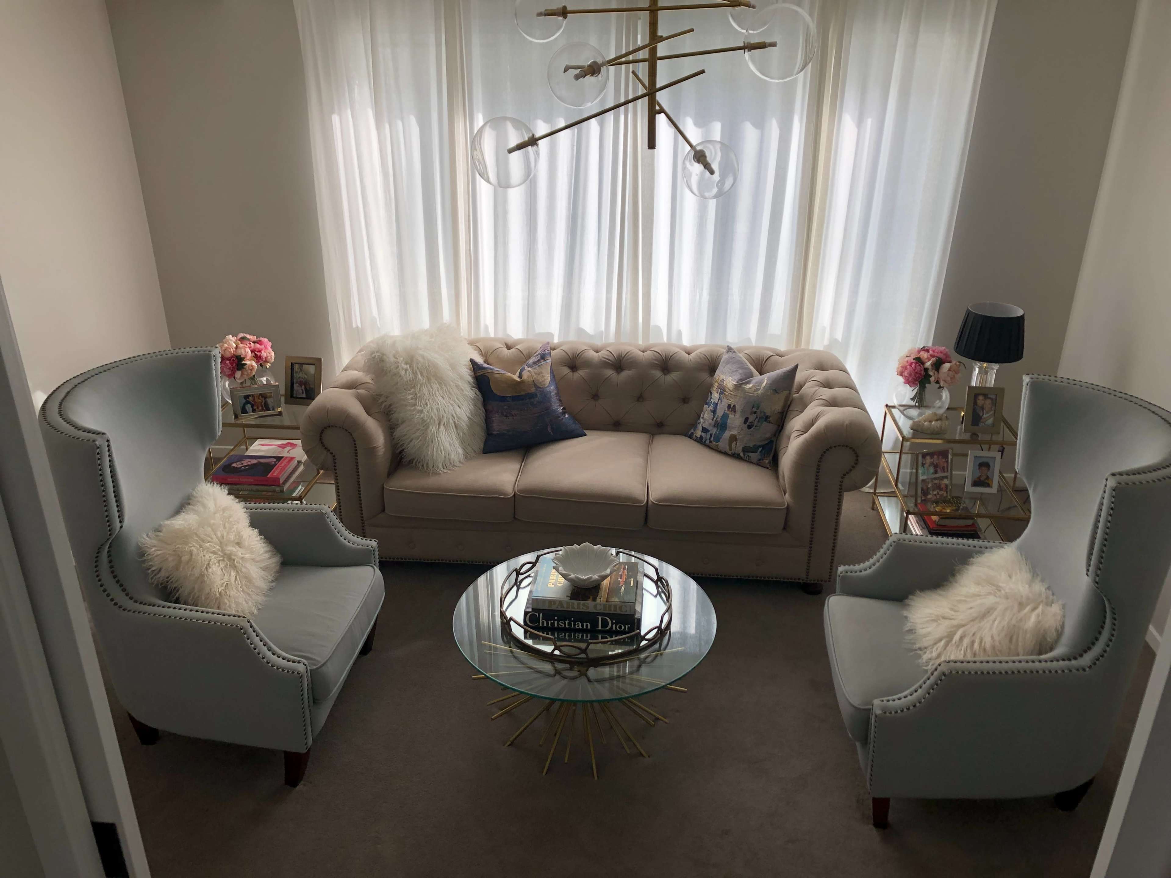 The image shows a cozy living room with a beige tufted sofa, two light blue chairs, a glass coffee table, and bright curtains.