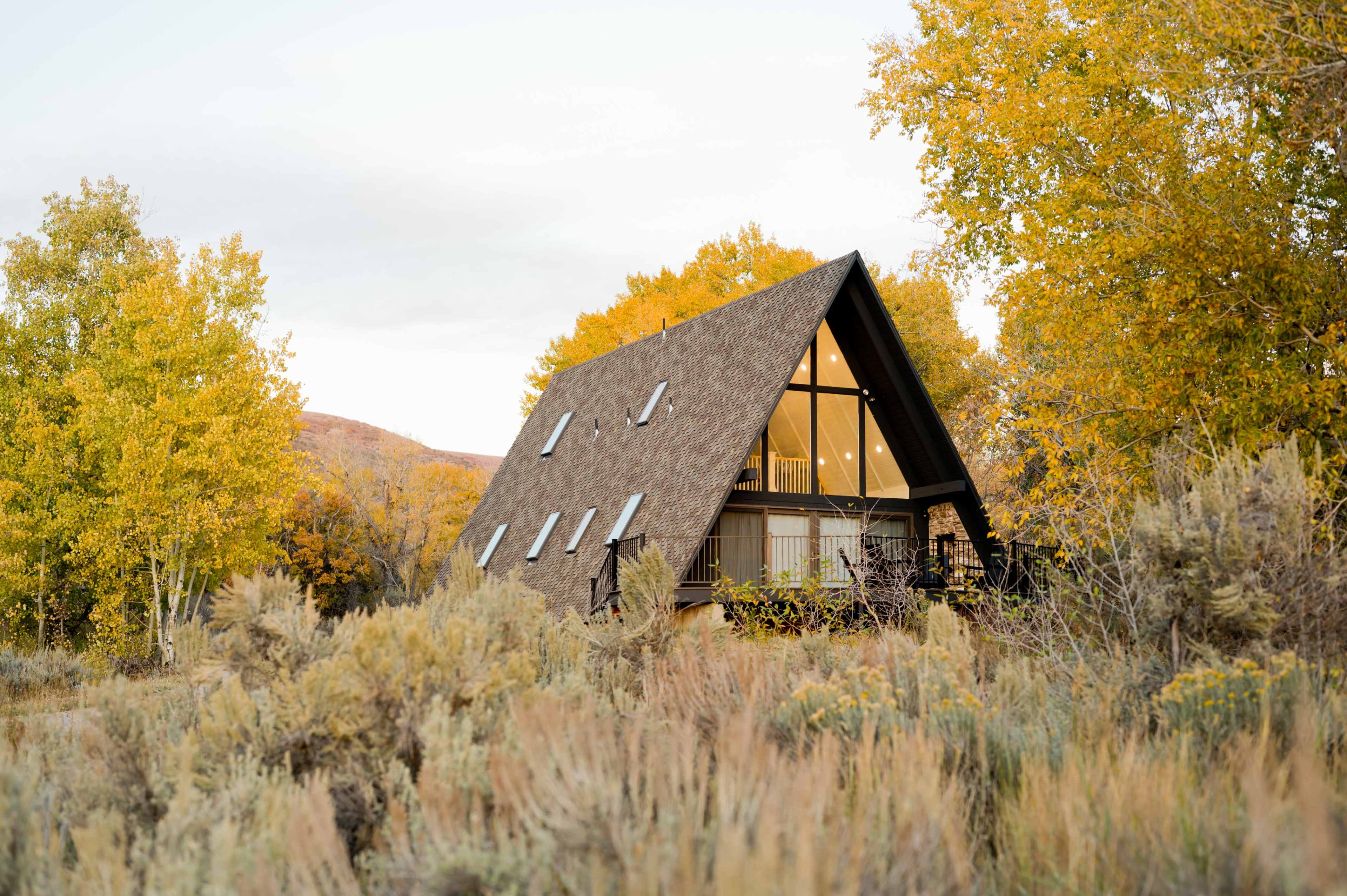 A triangular-roofed cabin is nestled among yellow-leaved trees and shrubs in a rural landscape.