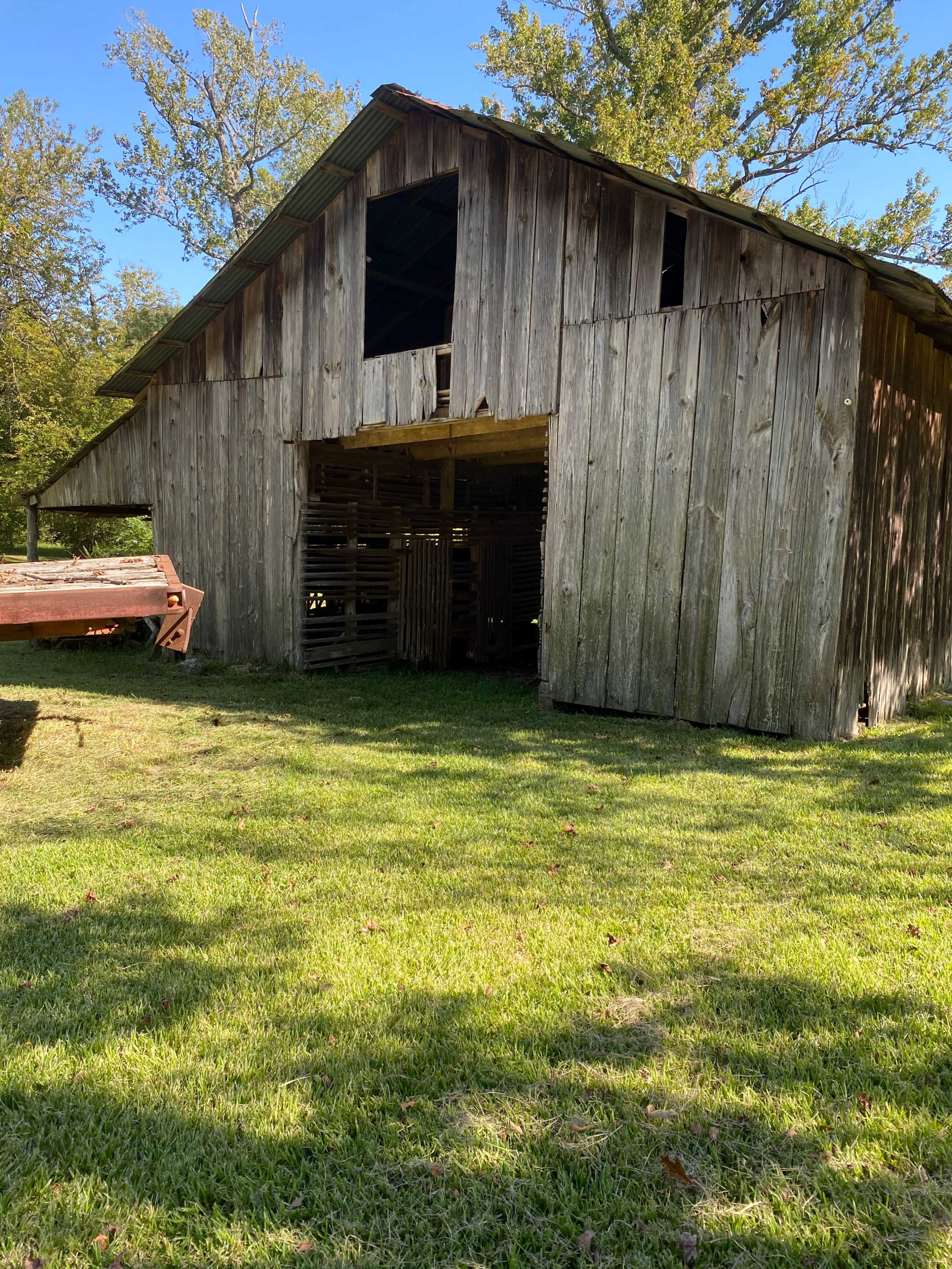 A weathered wooden barn with an open front, situated in a grassy area surrounded by trees.