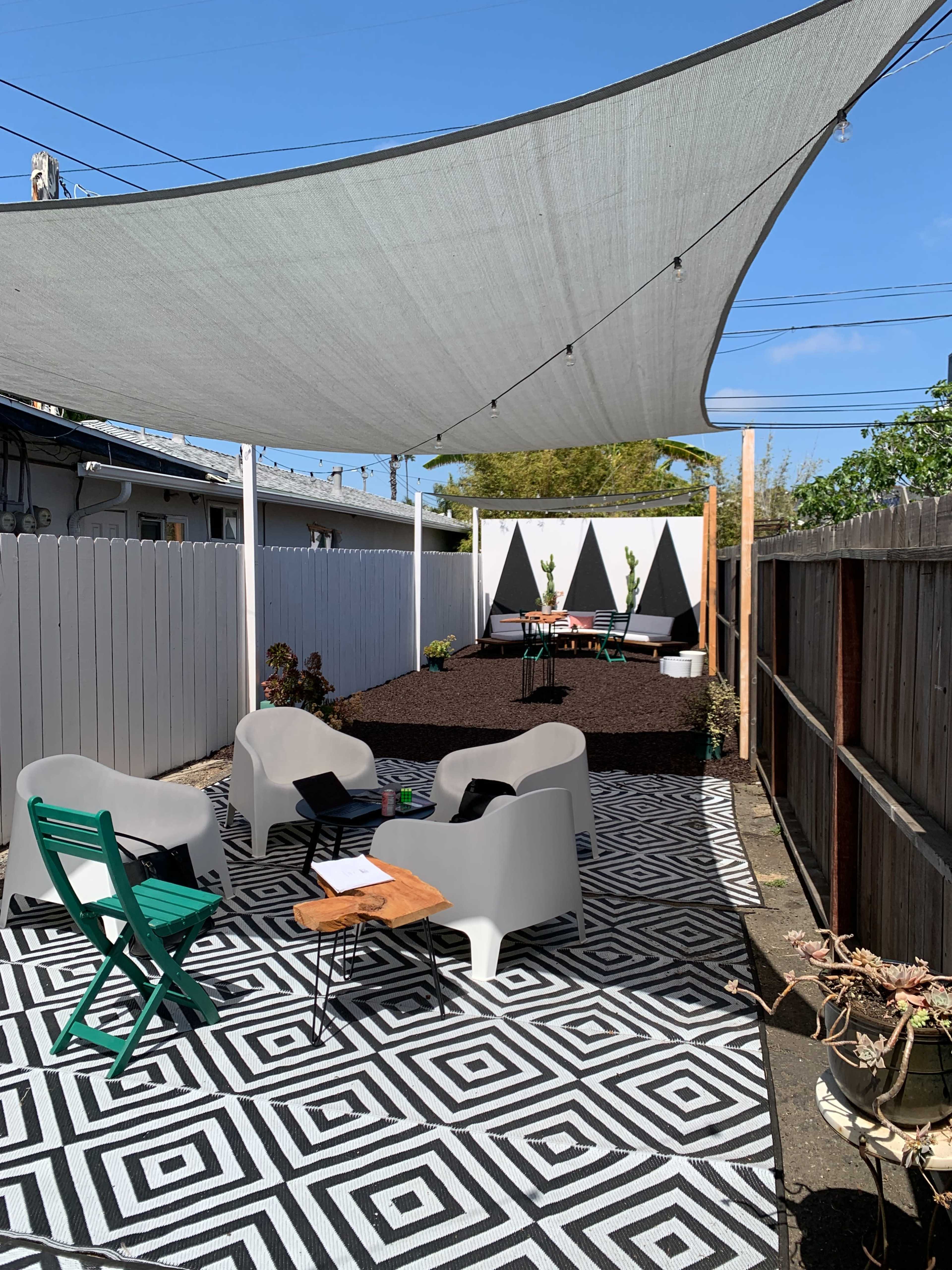 A patio area features a geometric patterned rug, several modern chairs, a table, and a large shade sail overhead, surrounded by a fence and greenery.