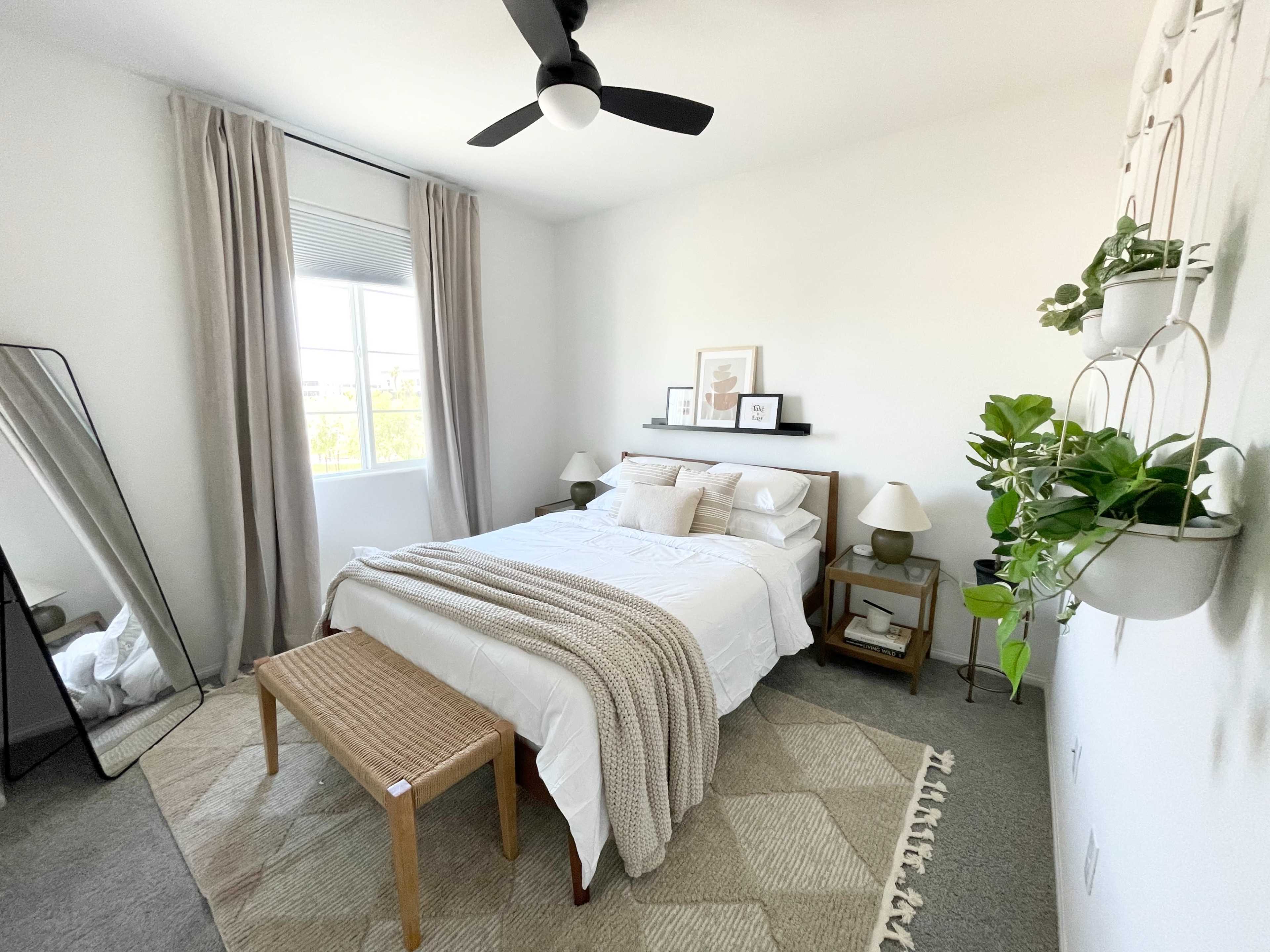 A neatly arranged bedroom features a bed with a light-colored blanket, a mirrored dresser, and decorative plants on the wall.