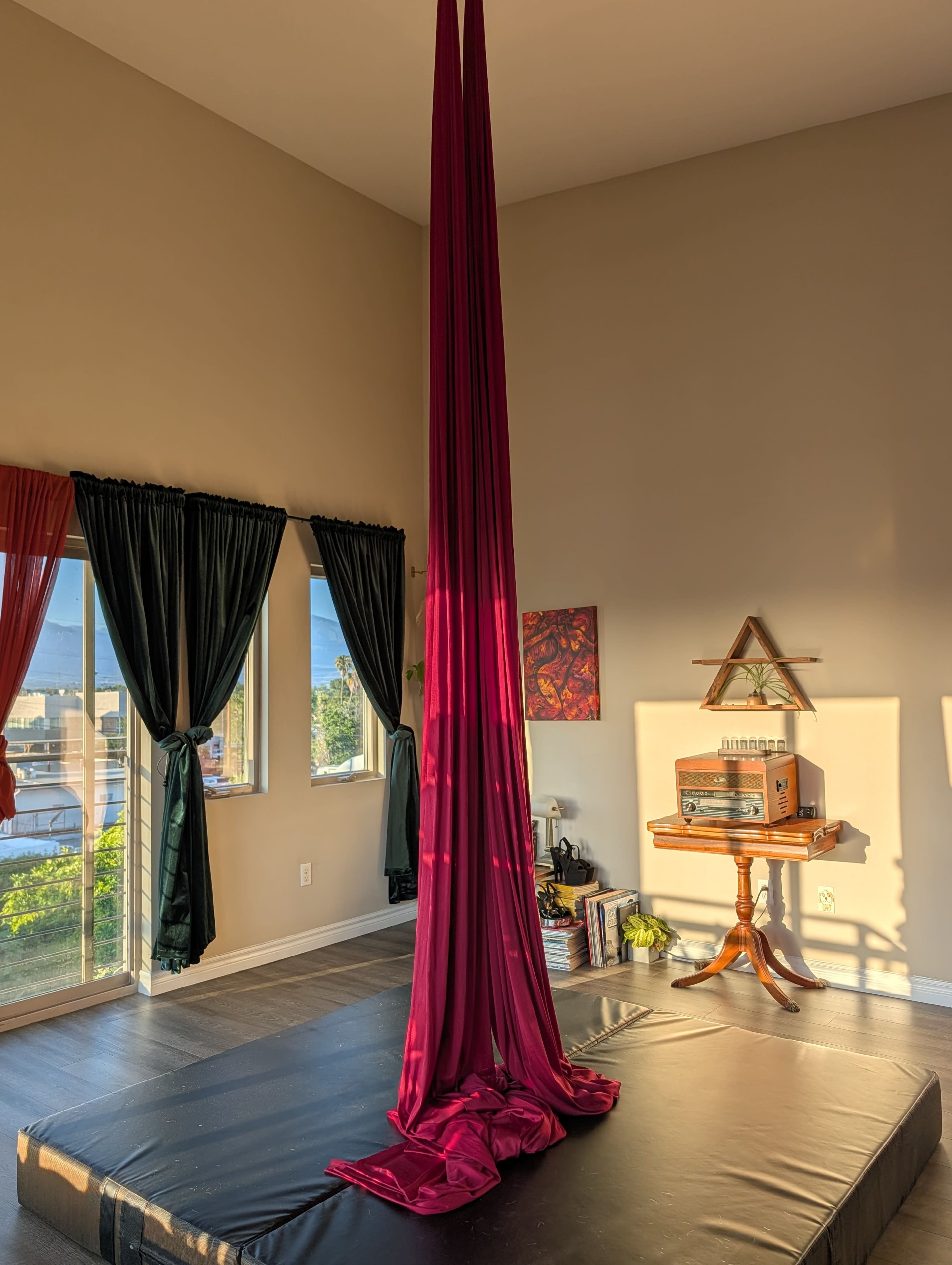 A maroon aerial silk hangs from the ceiling in a bright room with large windows and wooden furniture.