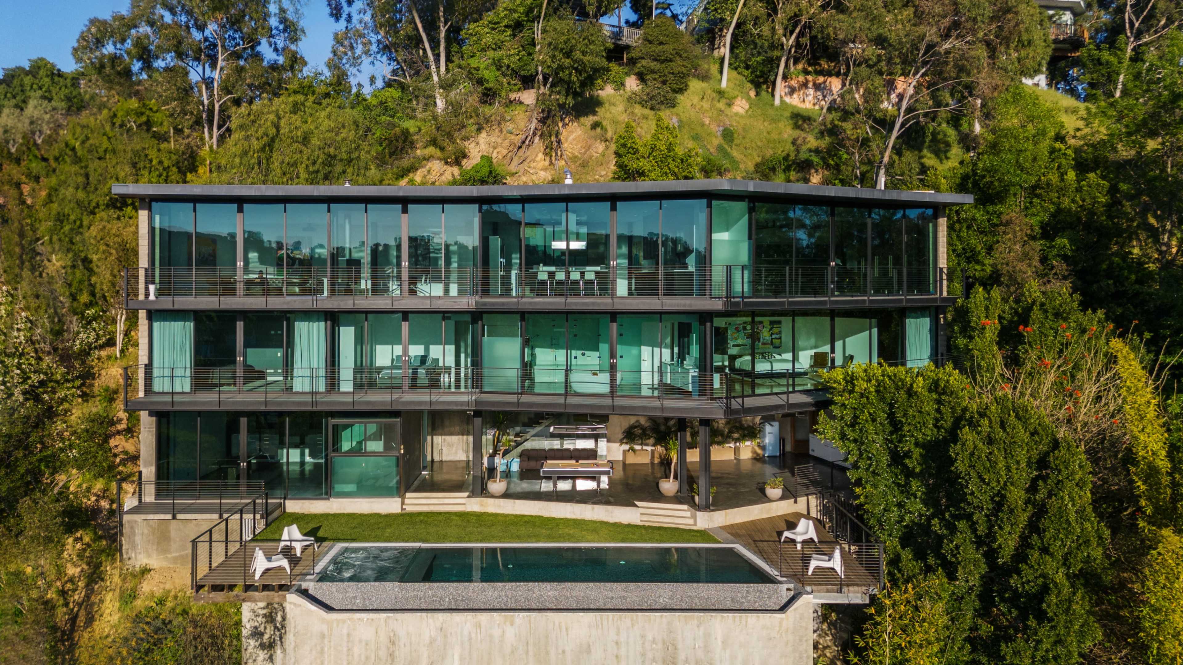 The image shows a modern glass-paneled house built on a hillside, featuring an outdoor pool and a terrace with seating areas.