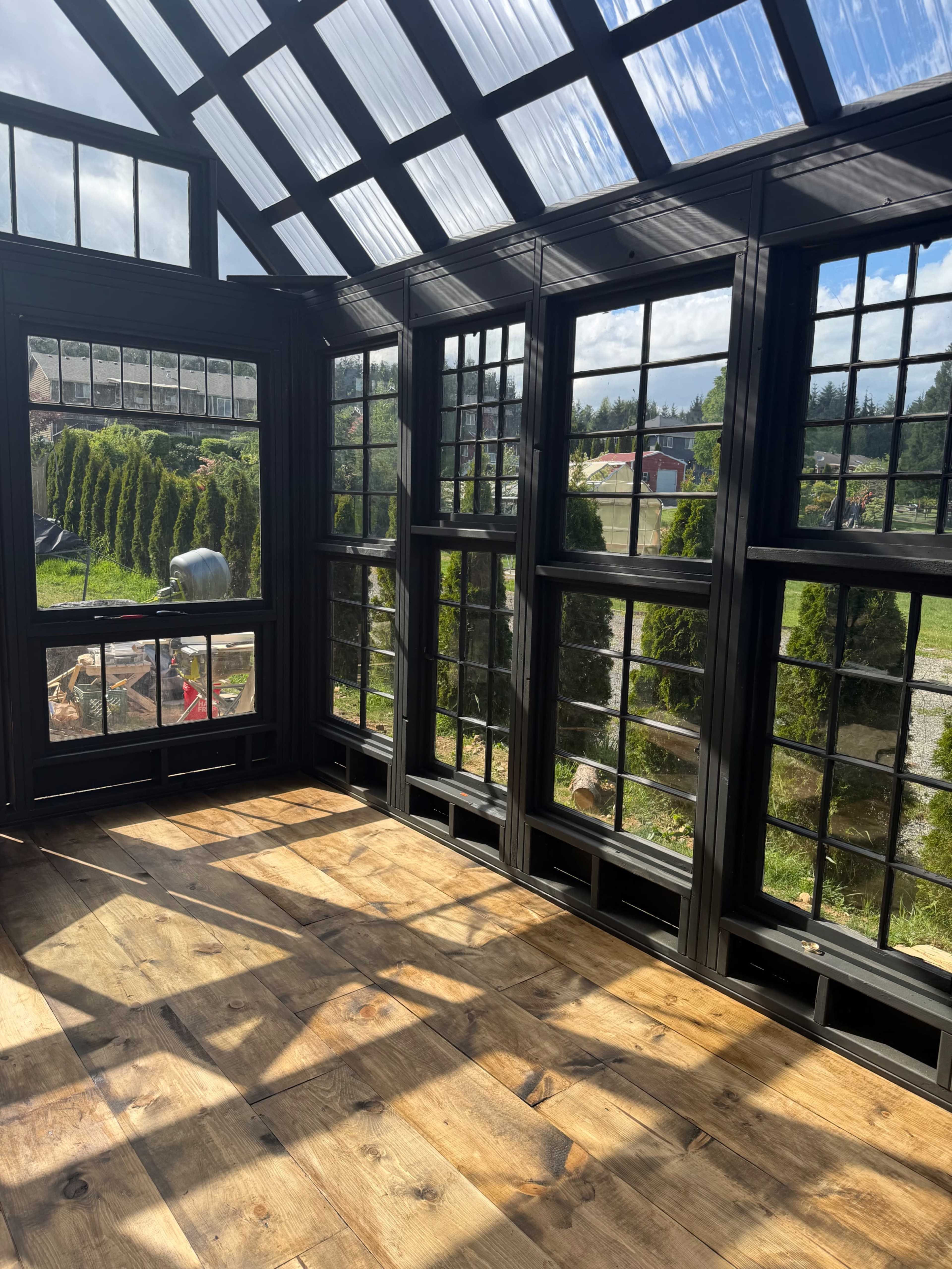 The image shows the interior of a greenhouse with large windows allowing natural light to enter and wooden flooring.