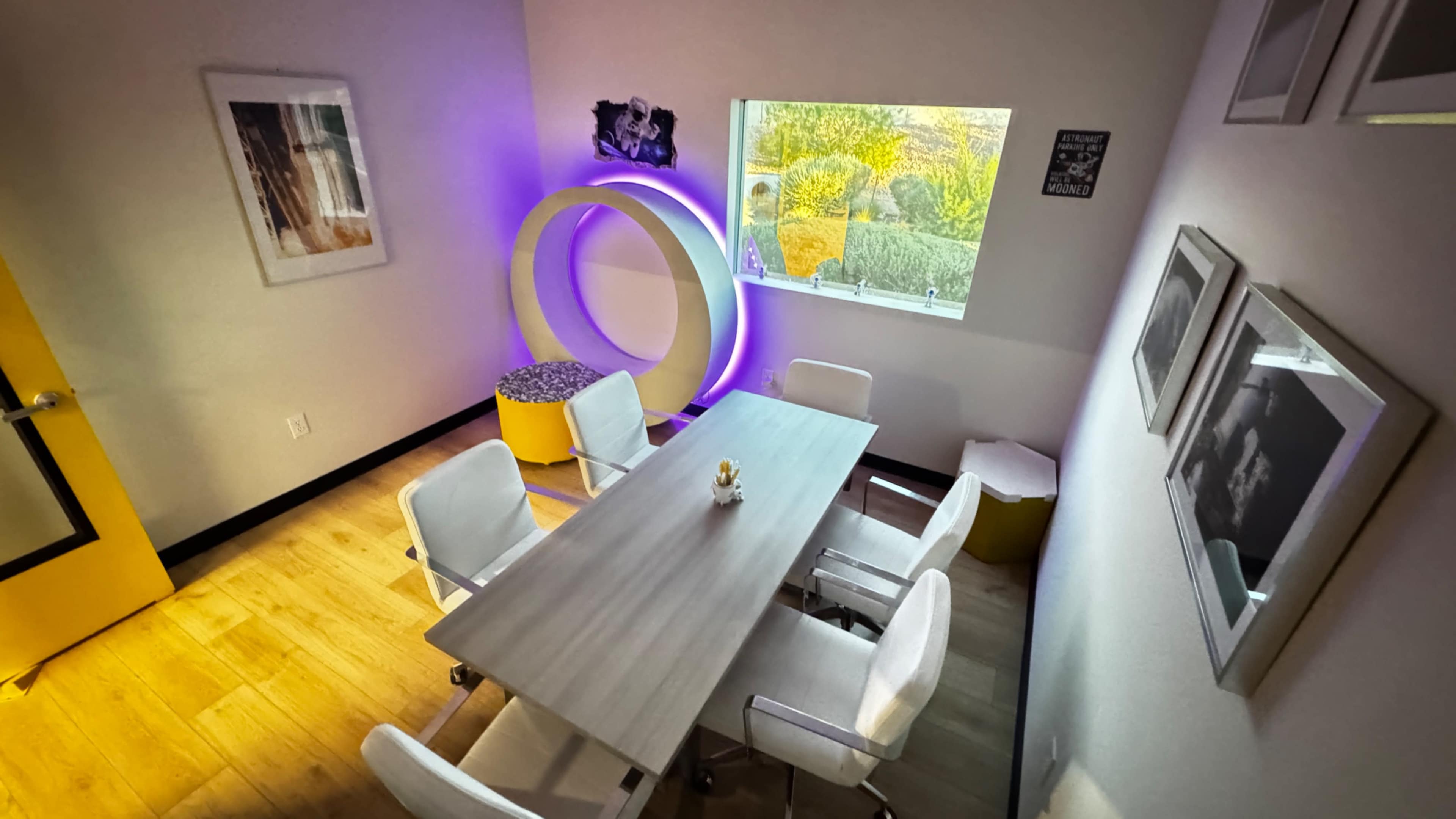 The image shows a modern meeting room featuring a rectangular table surrounded by white chairs, with a large circular decoration illuminated in purple and a window providing natural light.