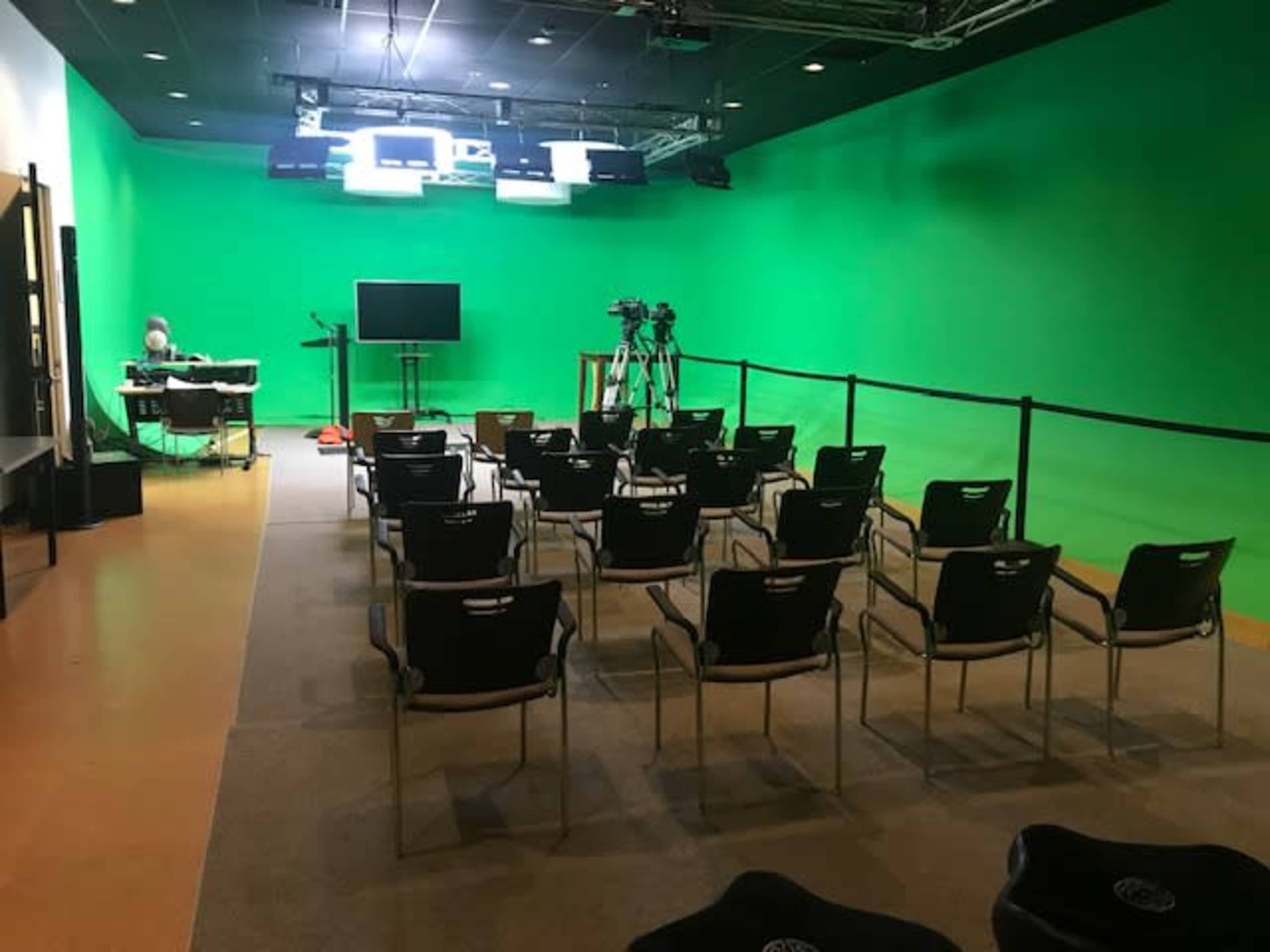 A green screen studio with rows of empty chairs facing a camera setup and an equipment table in the background.