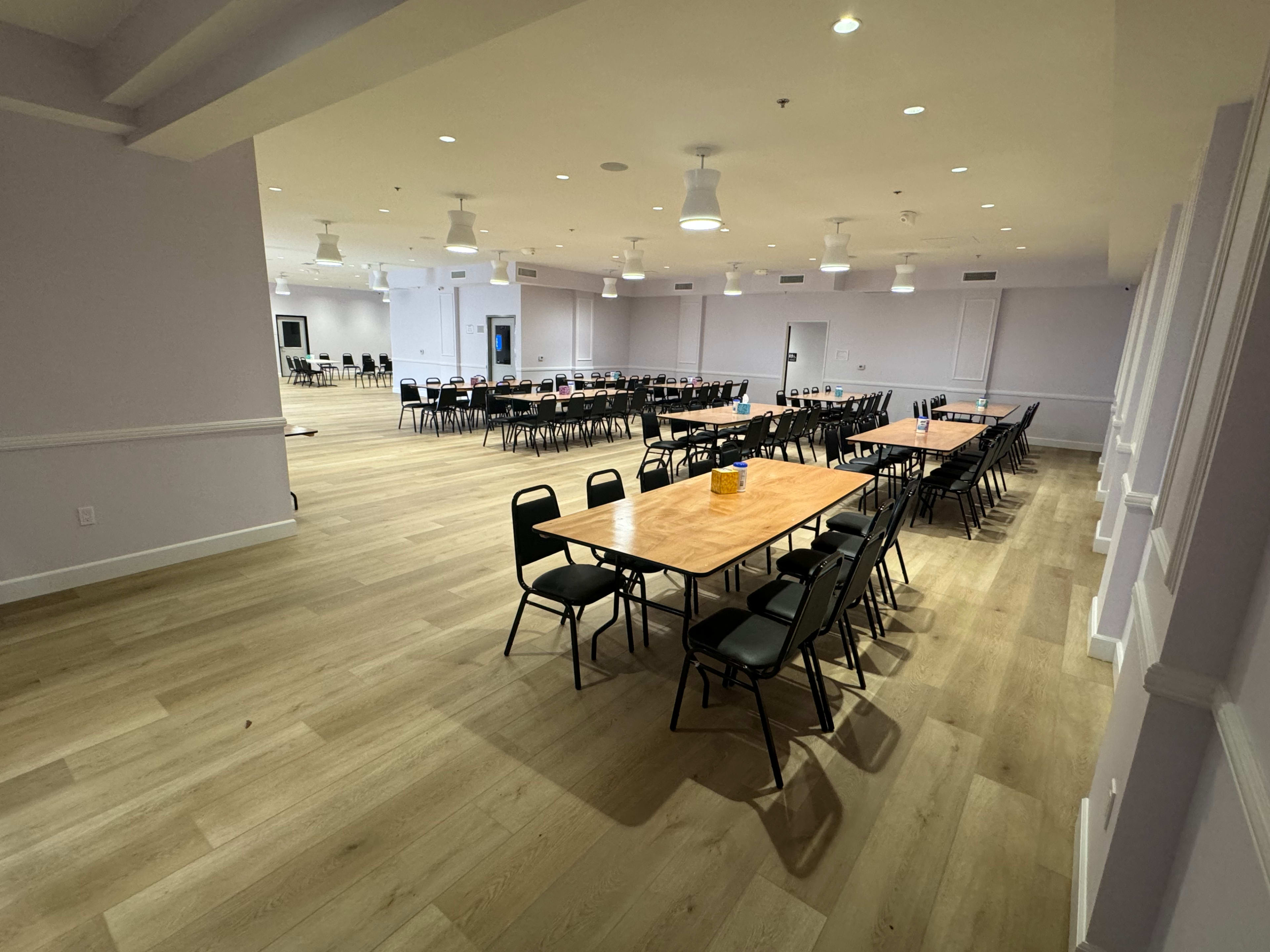 A spacious indoor venue with several rows of tables and chairs arranged in a modern layout, featuring wooden floors and bright overhead lighting.