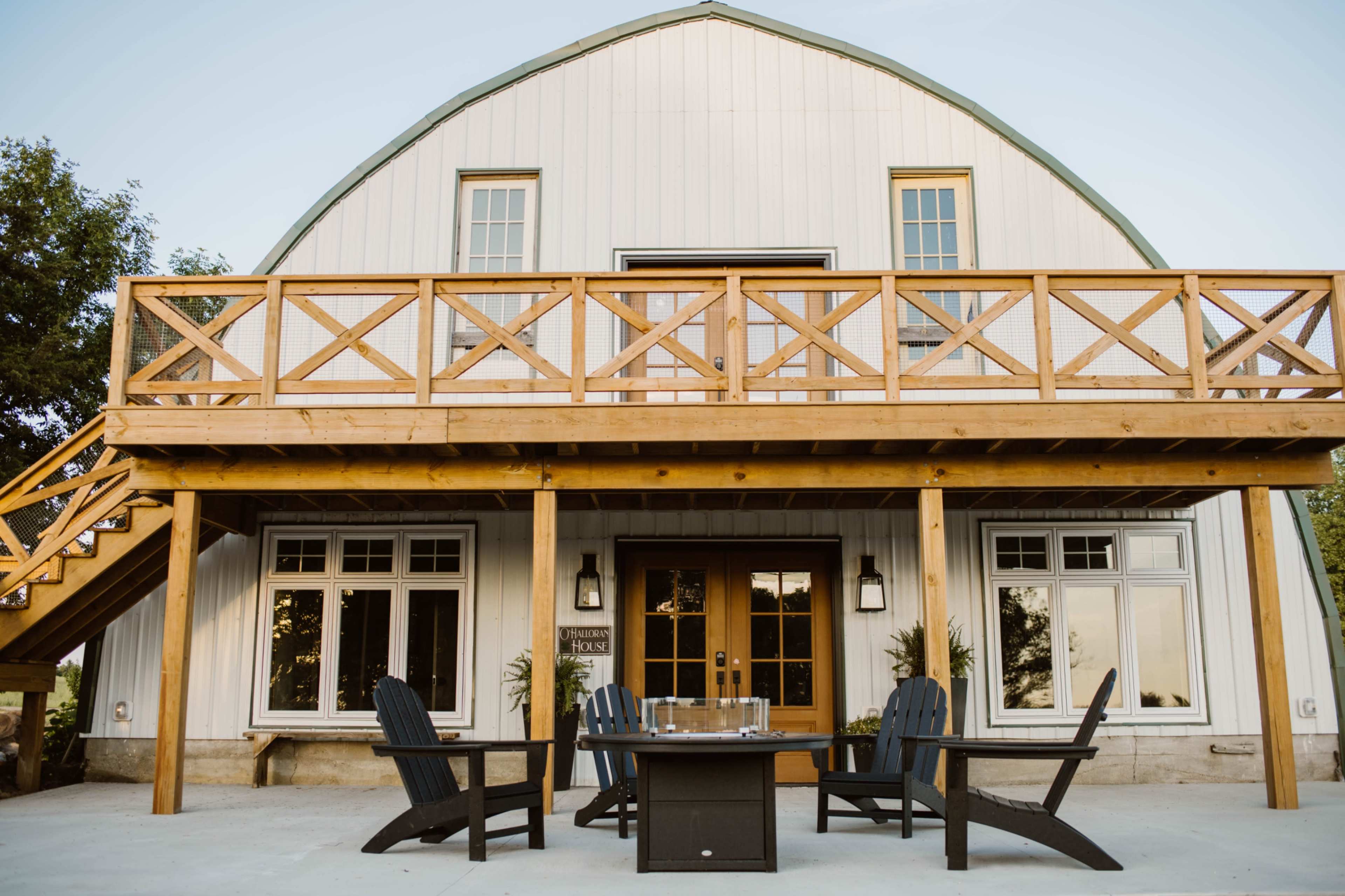 The image shows a large white barn-style building with a wooden deck and a seating area featuring four black chairs and a table in front.