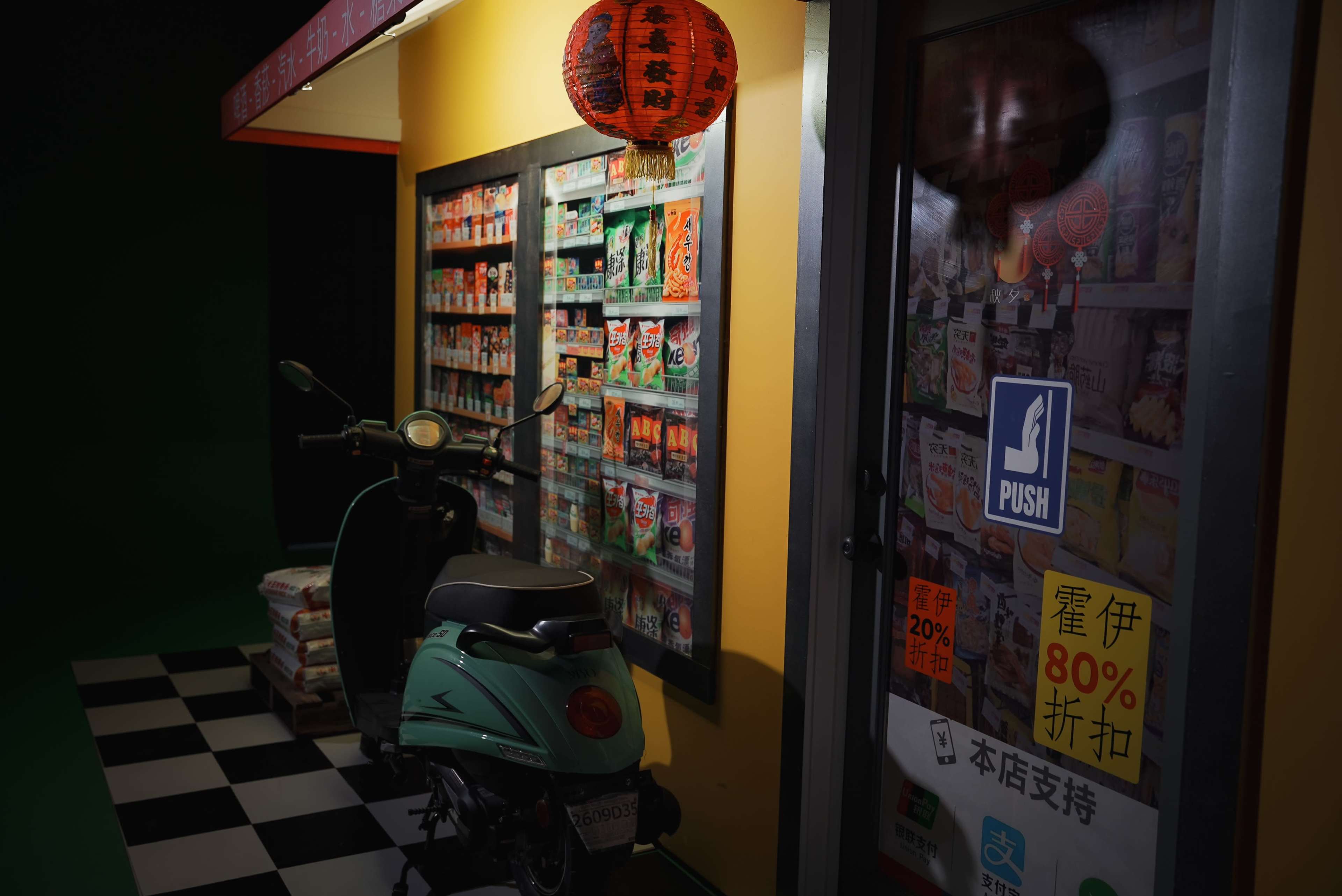 A colorful storefront displaying a variety of packaged goods, with a scooter parked beside the entrance and a lantern hanging above.