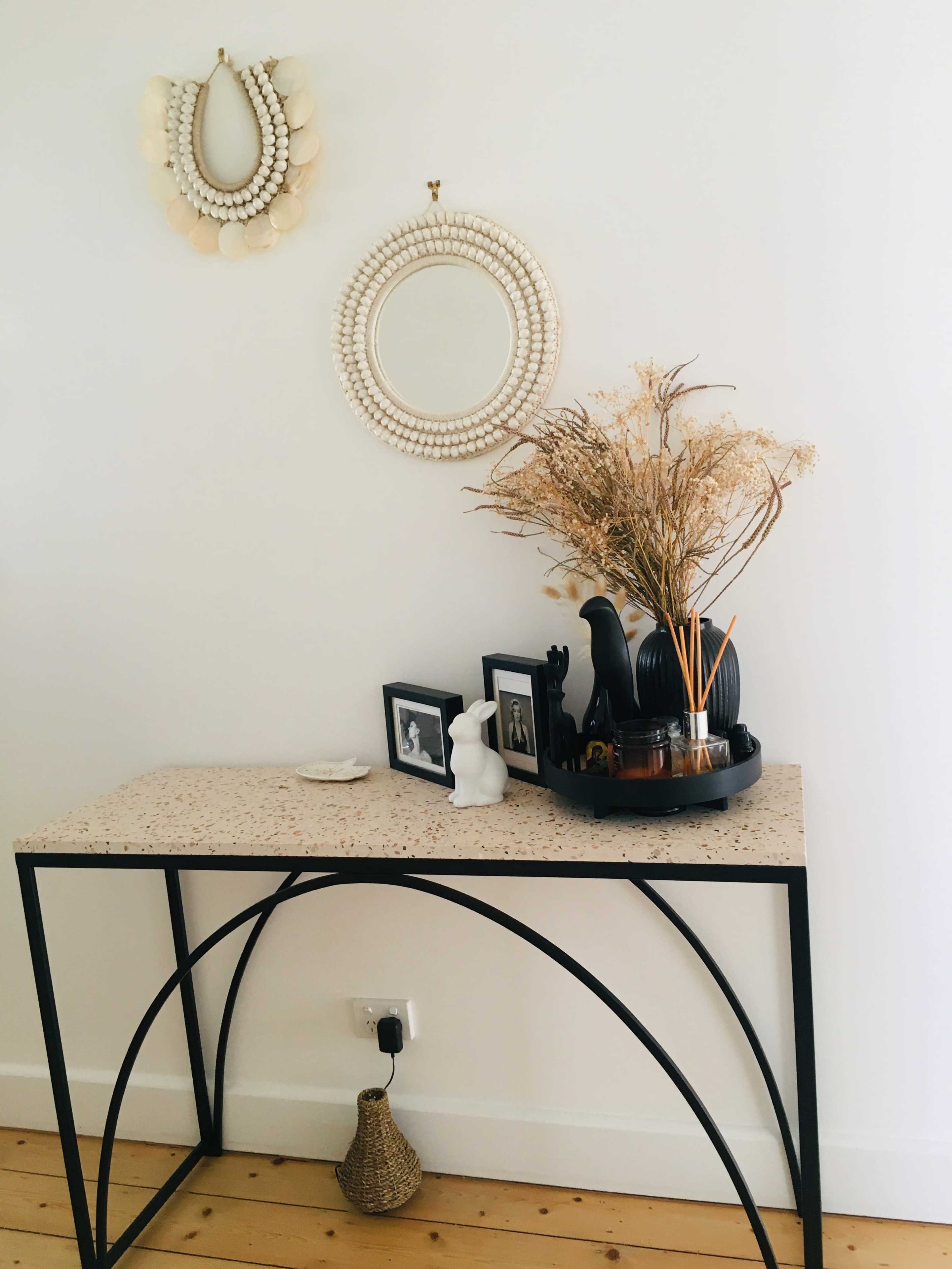 A console table with a speckled top displays a vase of dried flowers, framed photos, and decorative objects against a white wall adorned with a circular mirror and a hanging ornament.