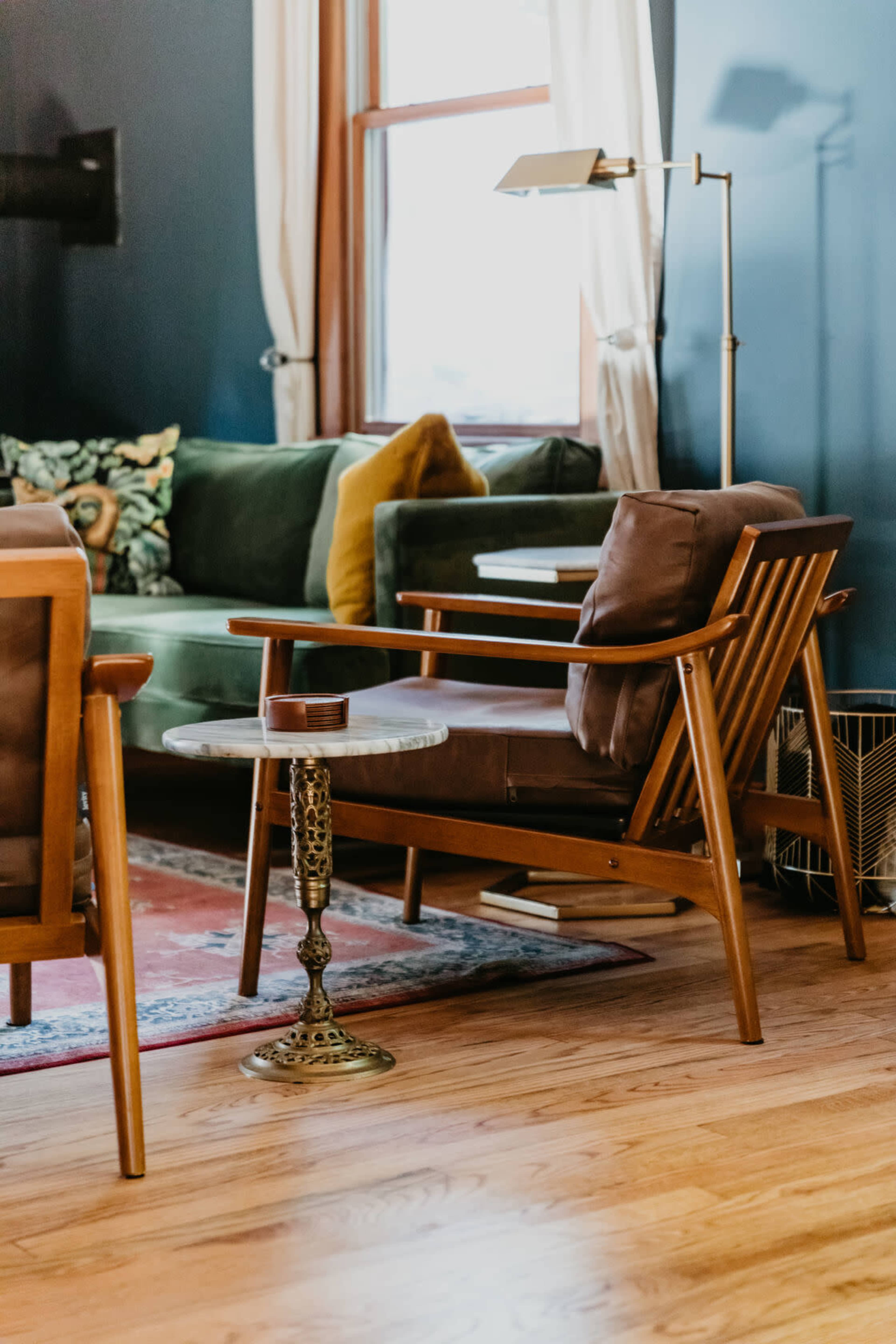 A wooden armchair with a small round side table sits adjacent to a green sofa in a well-lit living room.