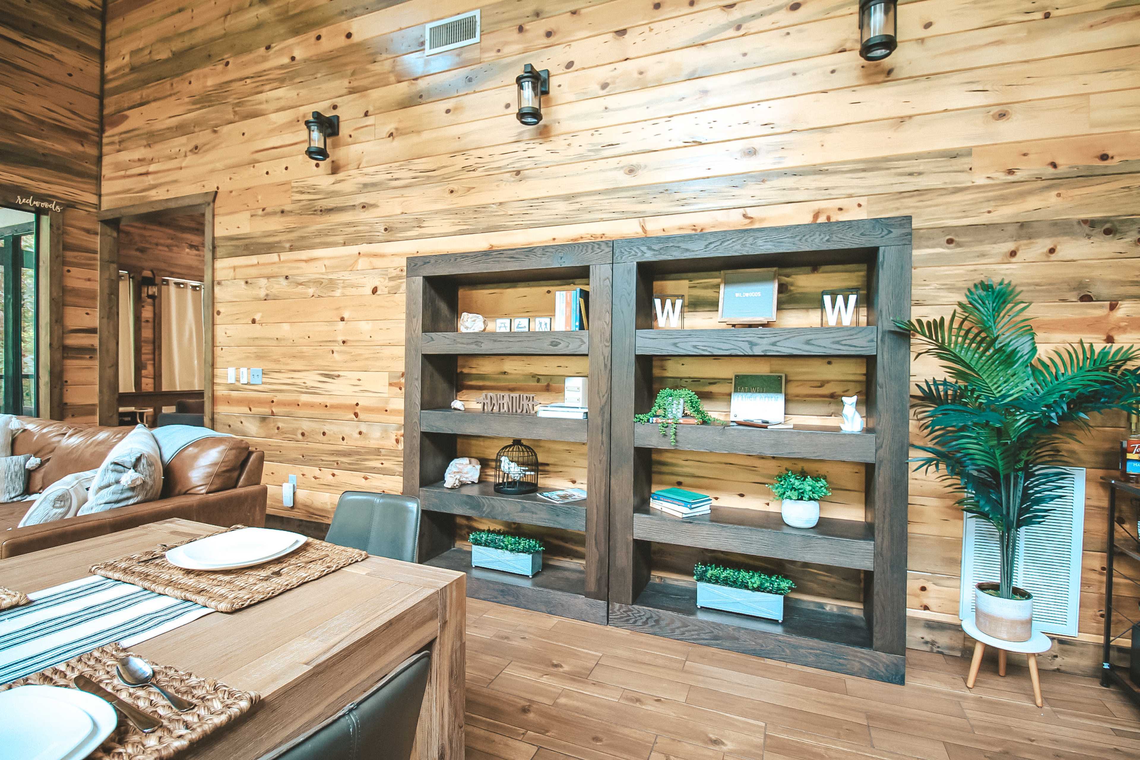 A wooden interior featuring two shelf units with decorative items, a dining table set, and a leafy plant in the corner.