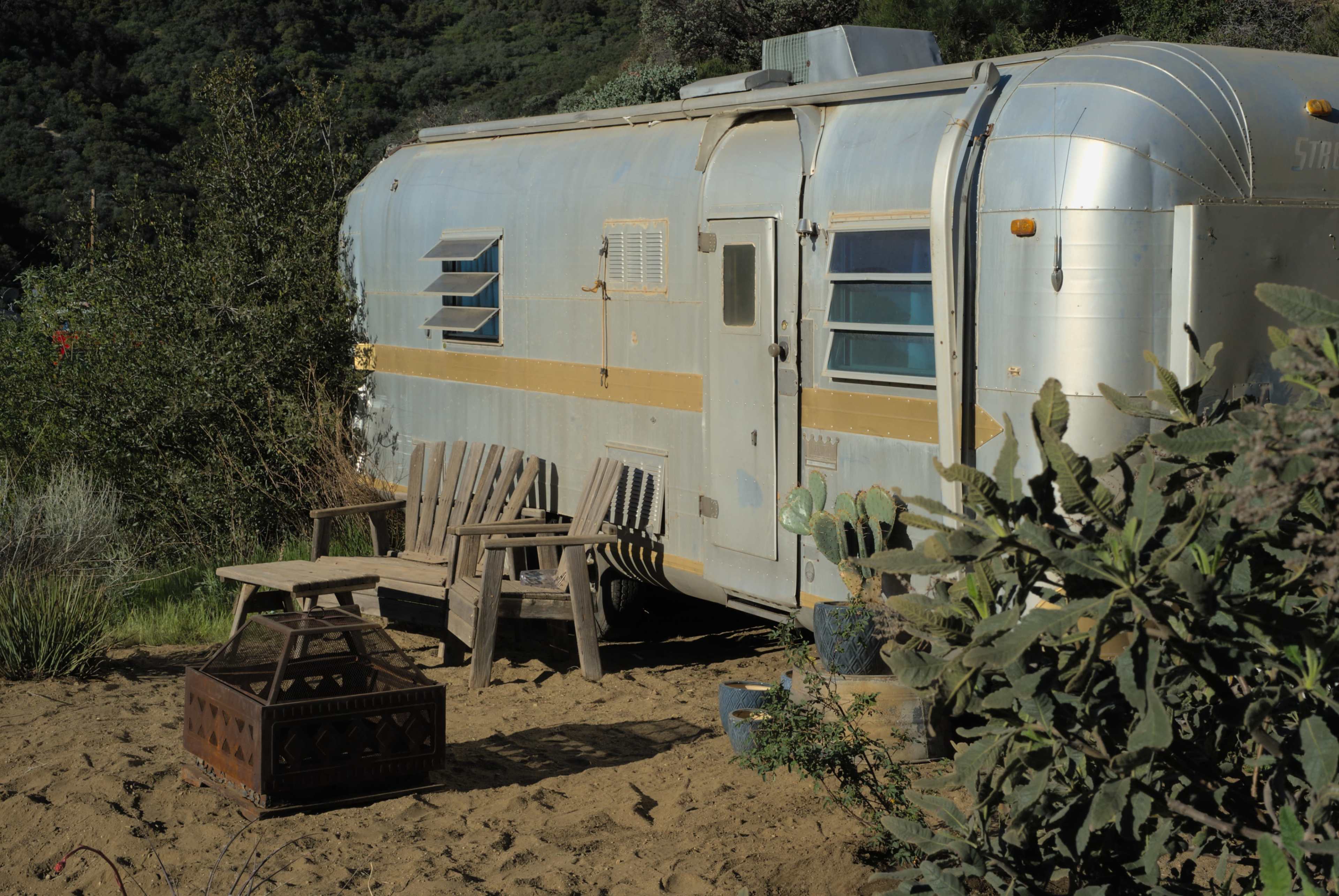 An aluminum retro trailer is parked on sandy ground surrounded by greenery, with two wooden chairs and a small table in front of it.