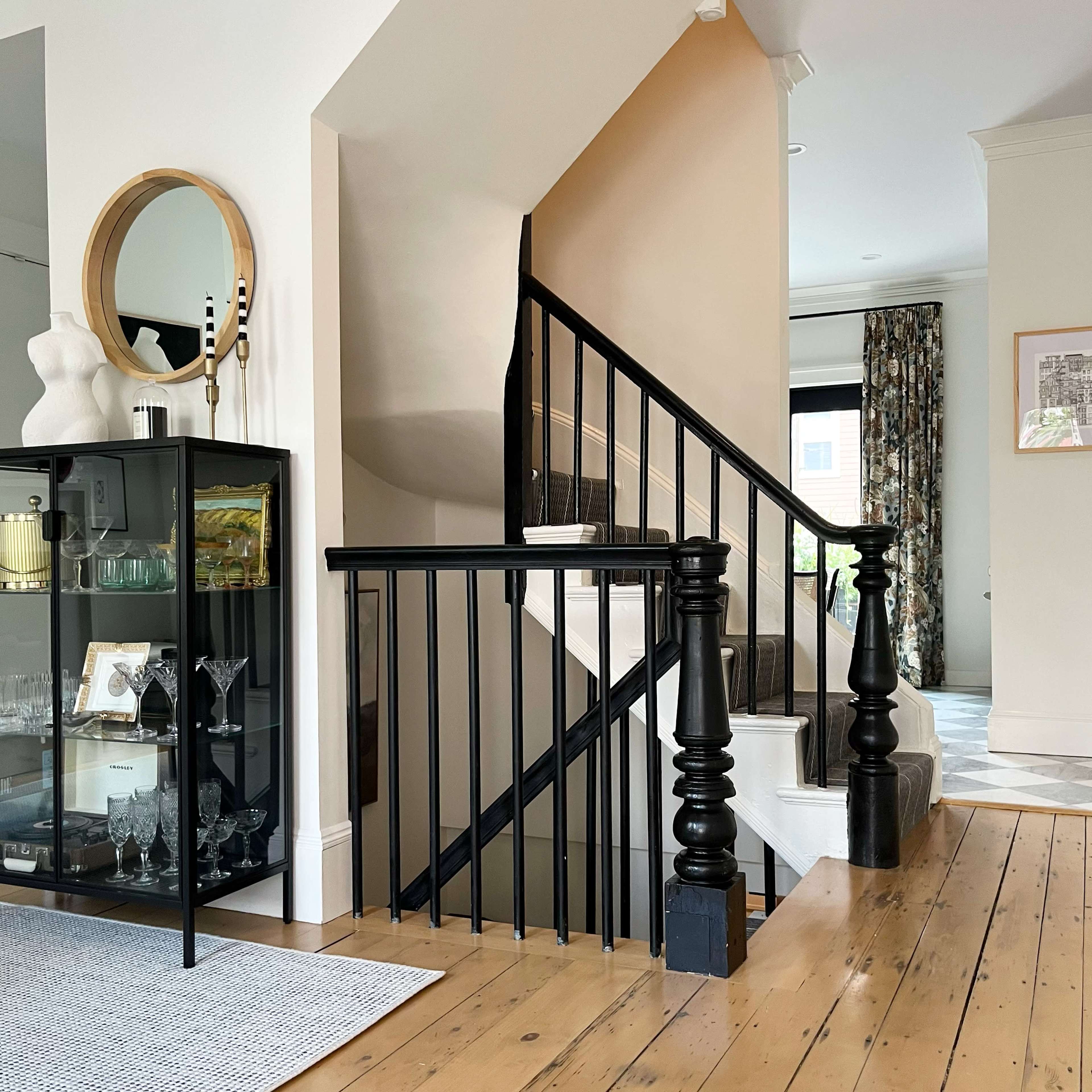 The image shows a modern interior featuring a staircase with black railings, light wooden floors, and a glass cabinet displaying glassware.