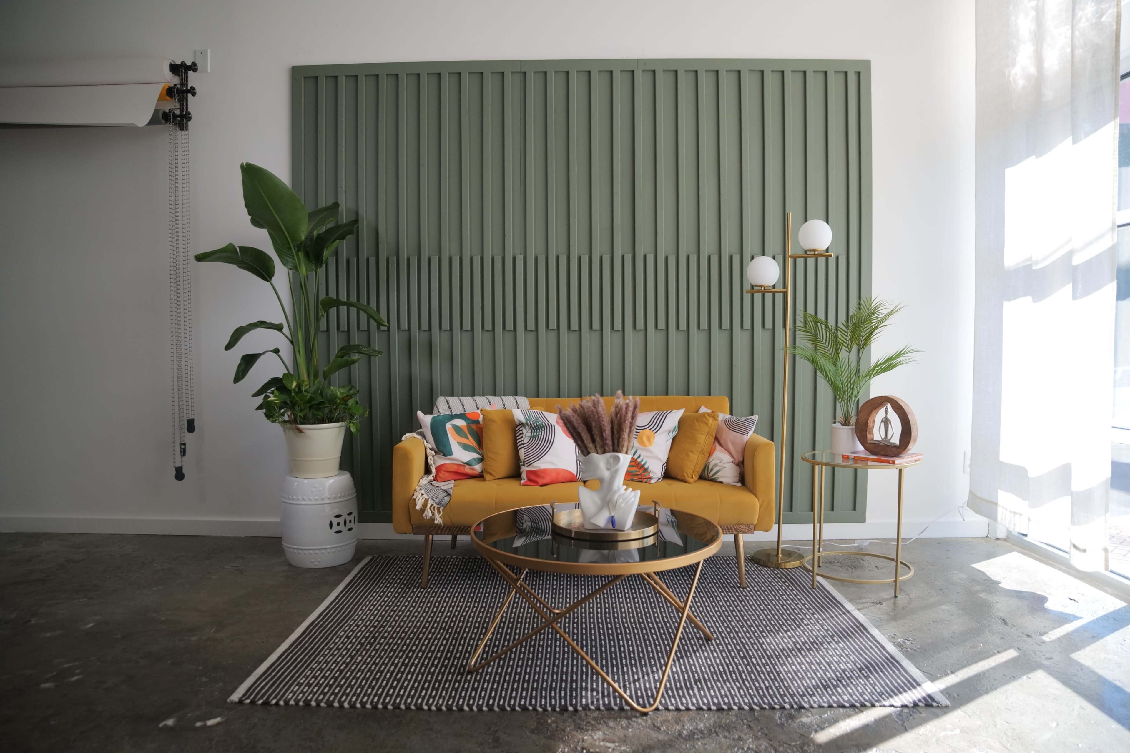 A modern living room features a mustard-colored sofa with decorative pillows, a round coffee table, and potted plants against a green paneled wall.