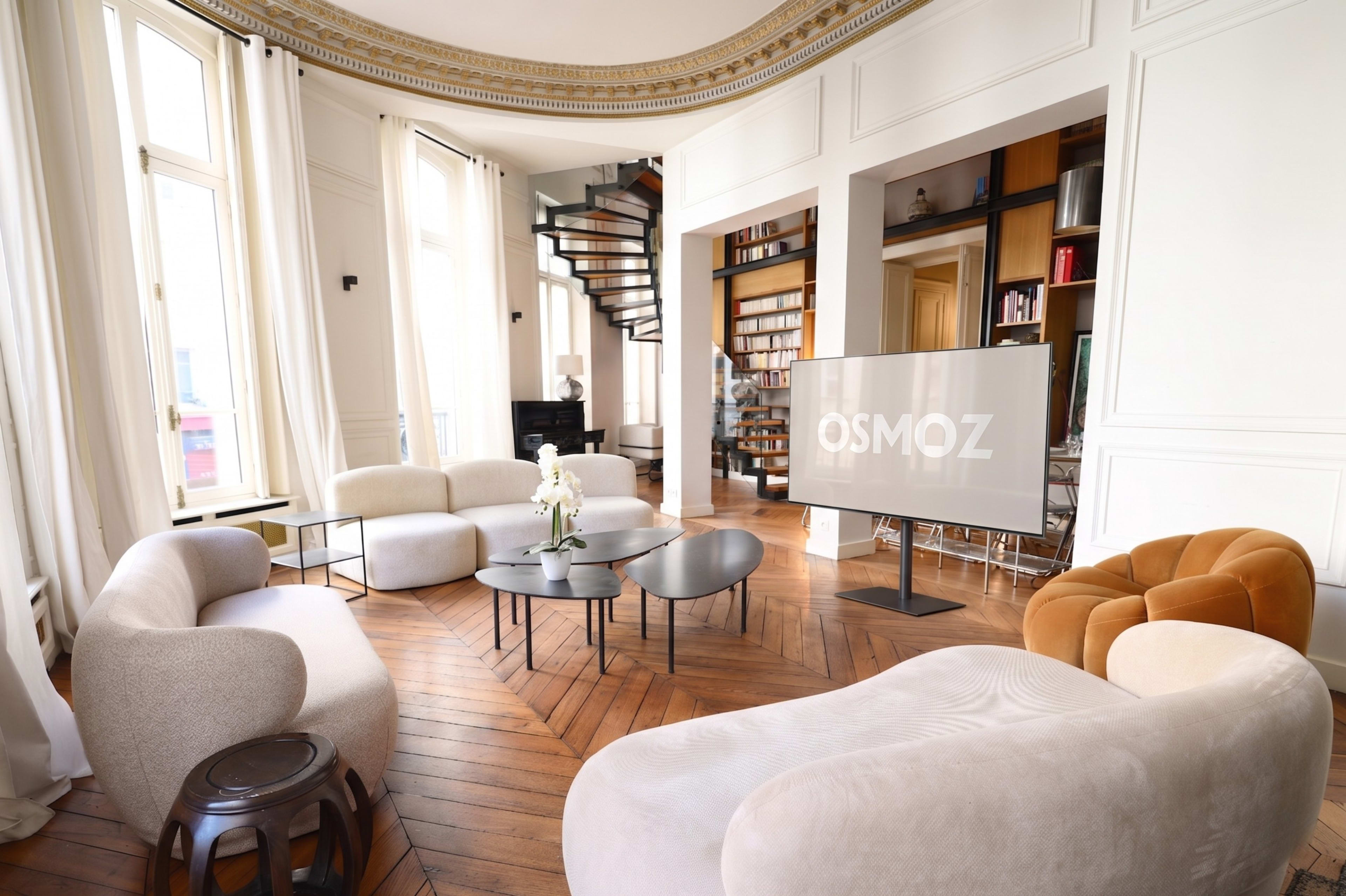 The image shows a modern living room with curved sofas, a television on a stand, and a wooden staircase in the background.
