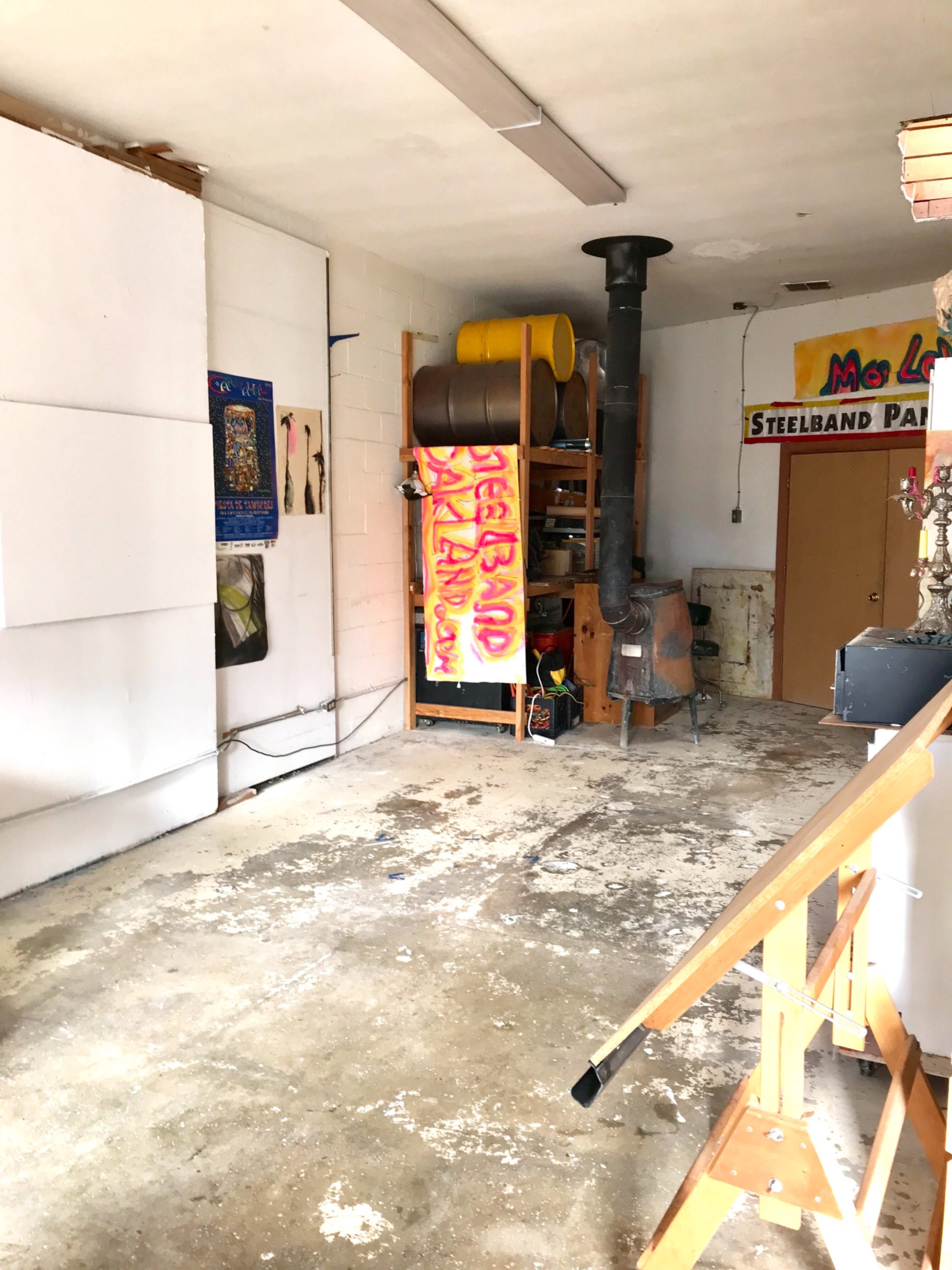 The image shows a partially empty garage or workshop with a concrete floor, shelving units, and a wood stove in the corner, along with colorful artwork hung on the walls.