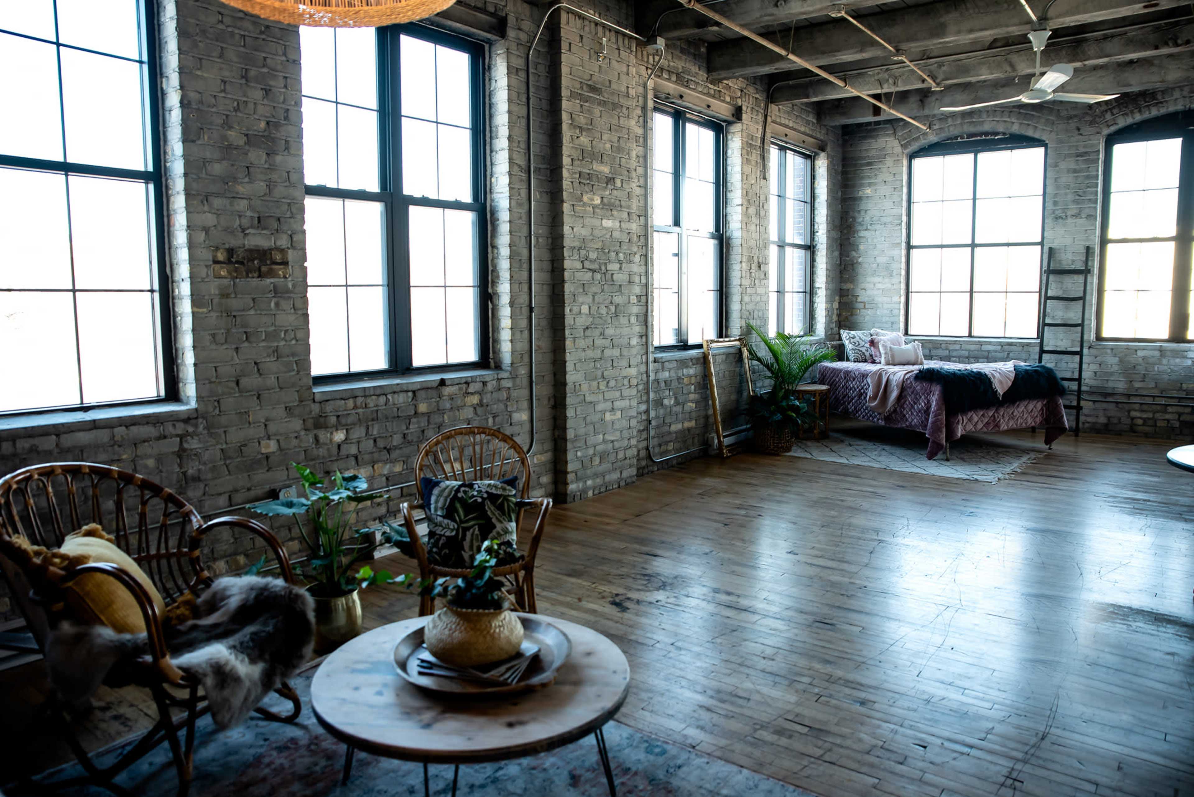 A spacious room features large windows, exposed brick walls, a bed dressed in patterned linens, and a seating area with wicker chairs and a round table.