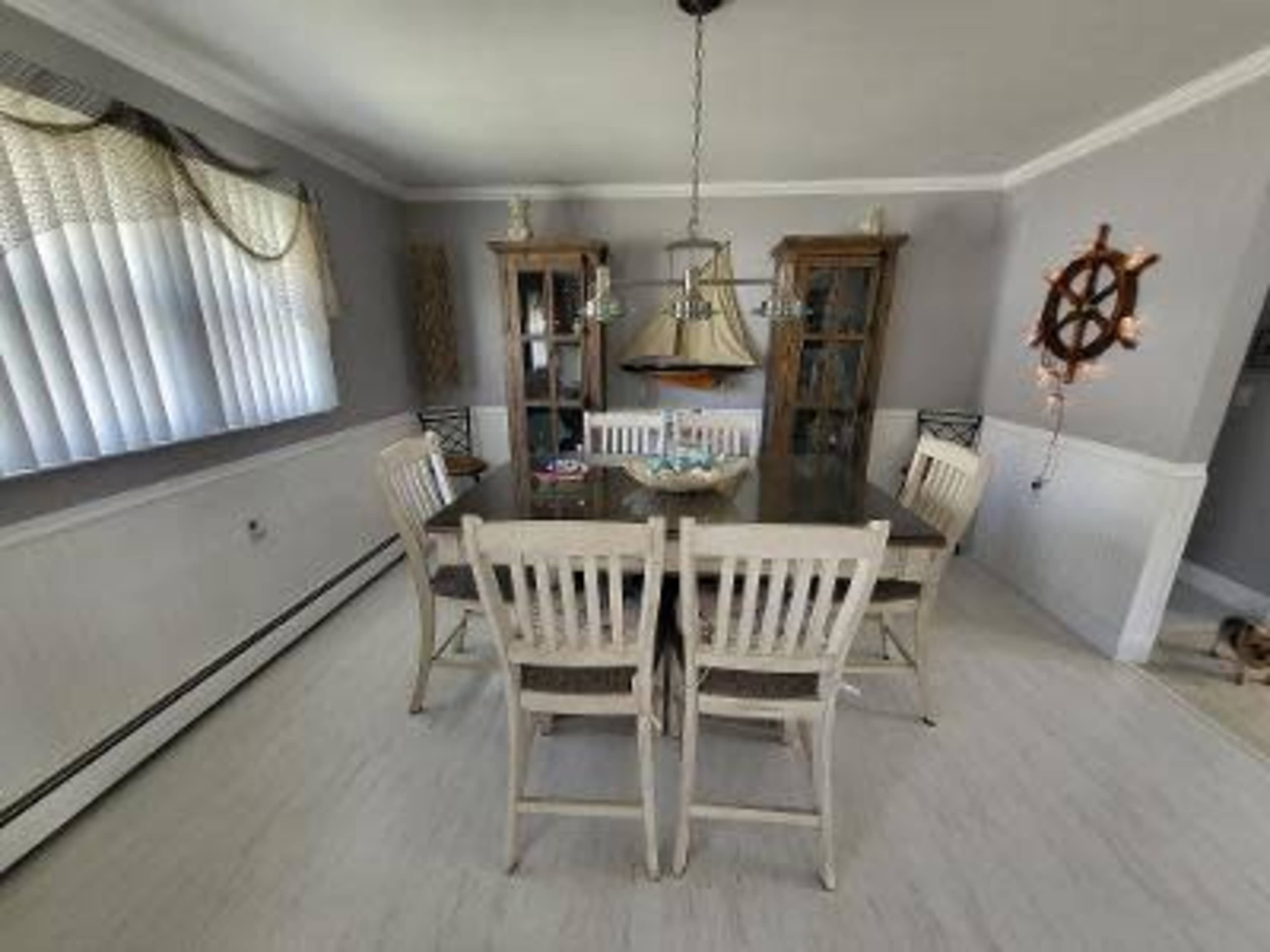 A dining room features a large table surrounded by six chairs, with two wooden cabinets displaying items and a light fixture above.