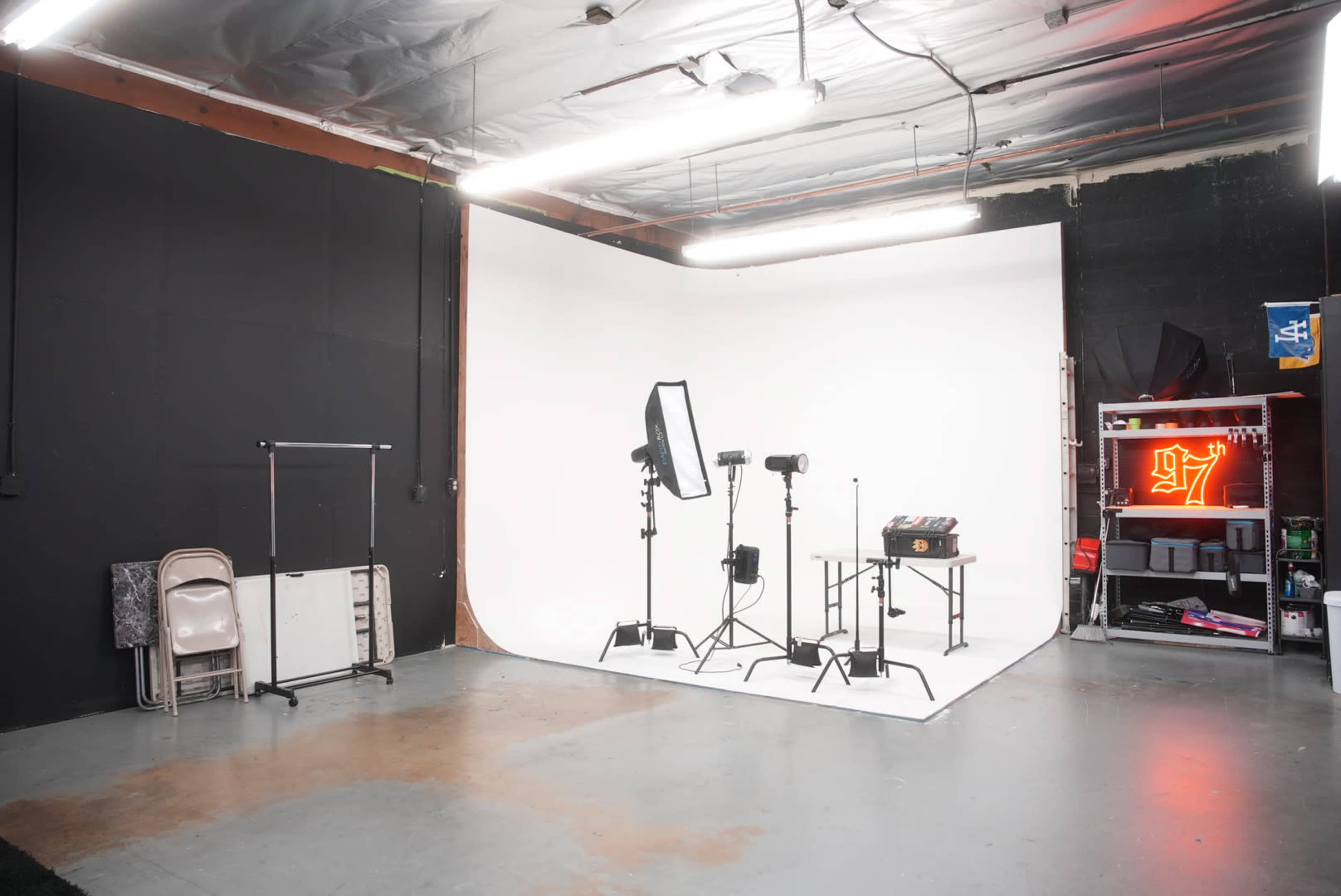 A photography studio featuring a white backdrop, lighting equipment, a table, and a storage area with various items.
