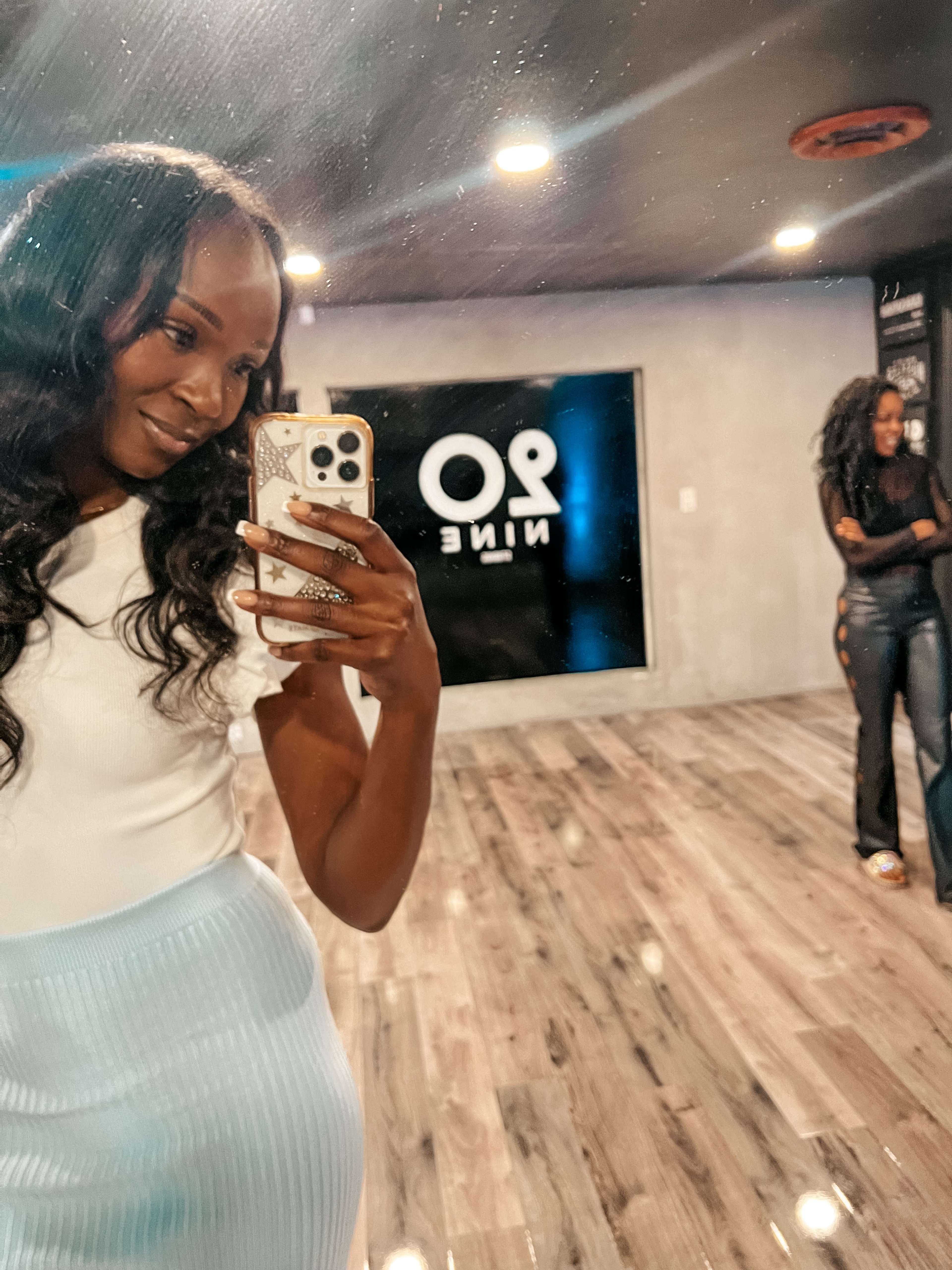 A woman takes a selfie in a mirror while another woman stands in the background.
