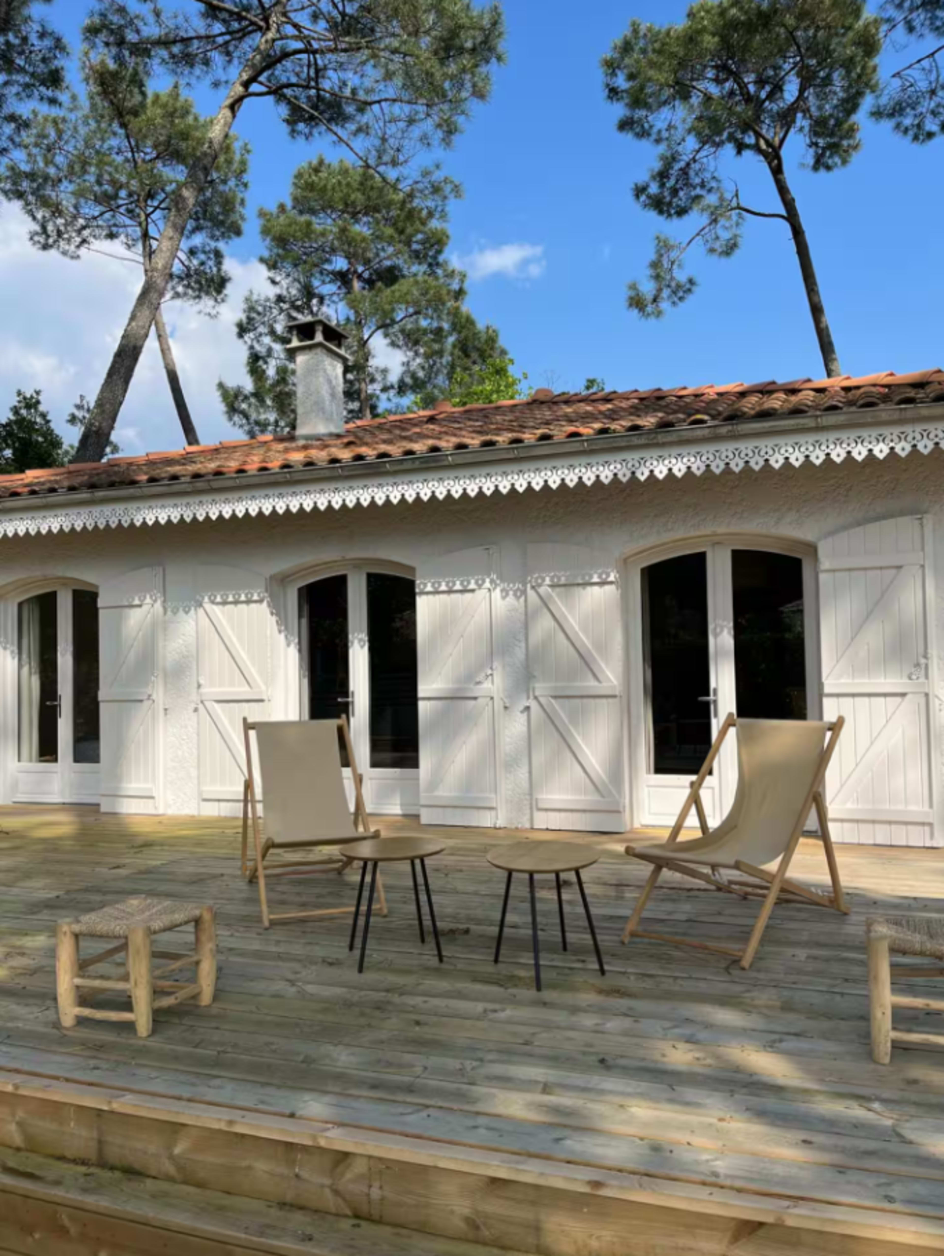 A wooden deck features two chairs and a small table in front of a house with white shutters and a tiled roof, surrounded by tall trees under a clear blue sky.