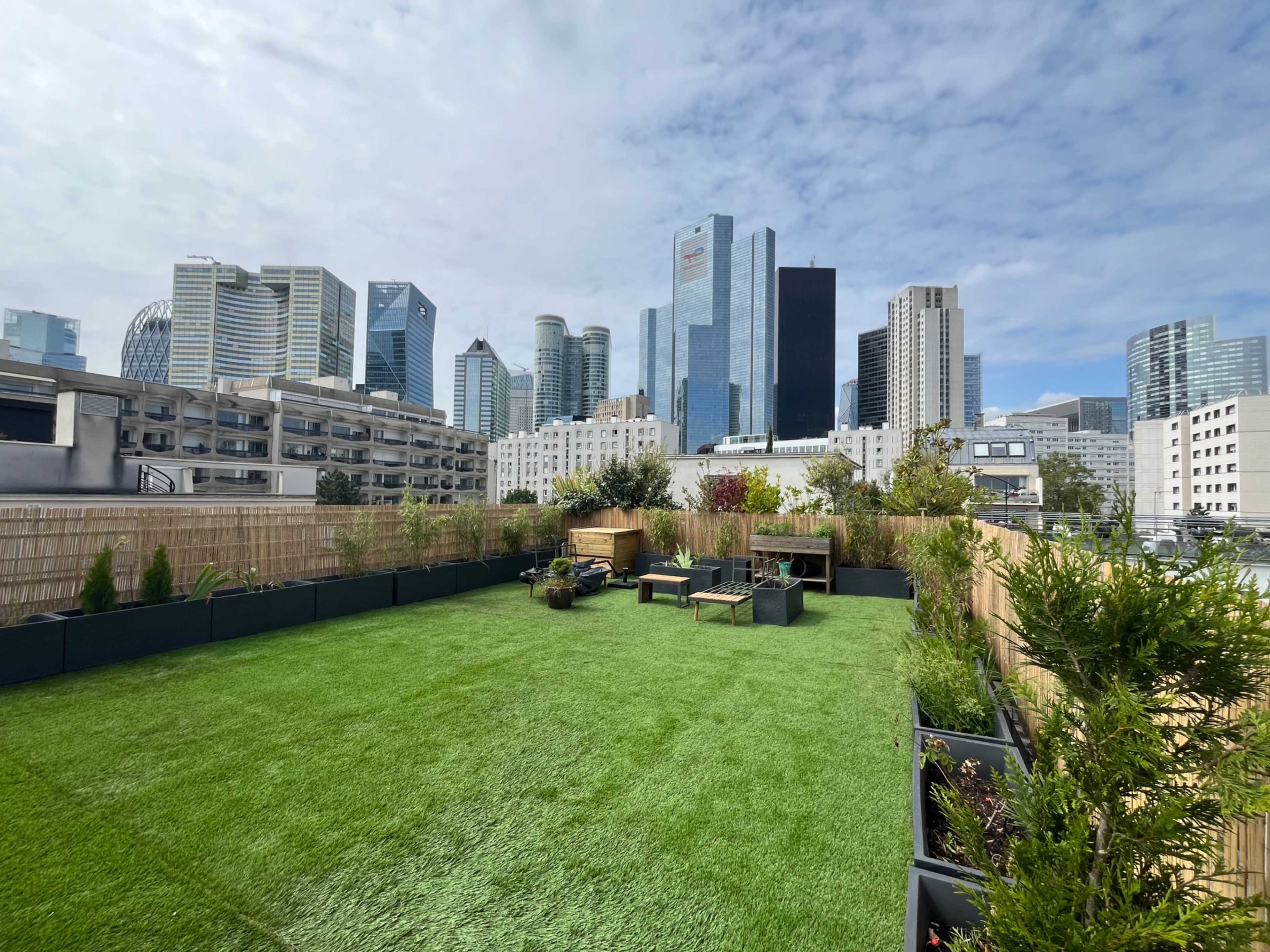 A rooftop terrace features artificial grass and seating, surrounded by modern skyscrapers in an urban landscape.