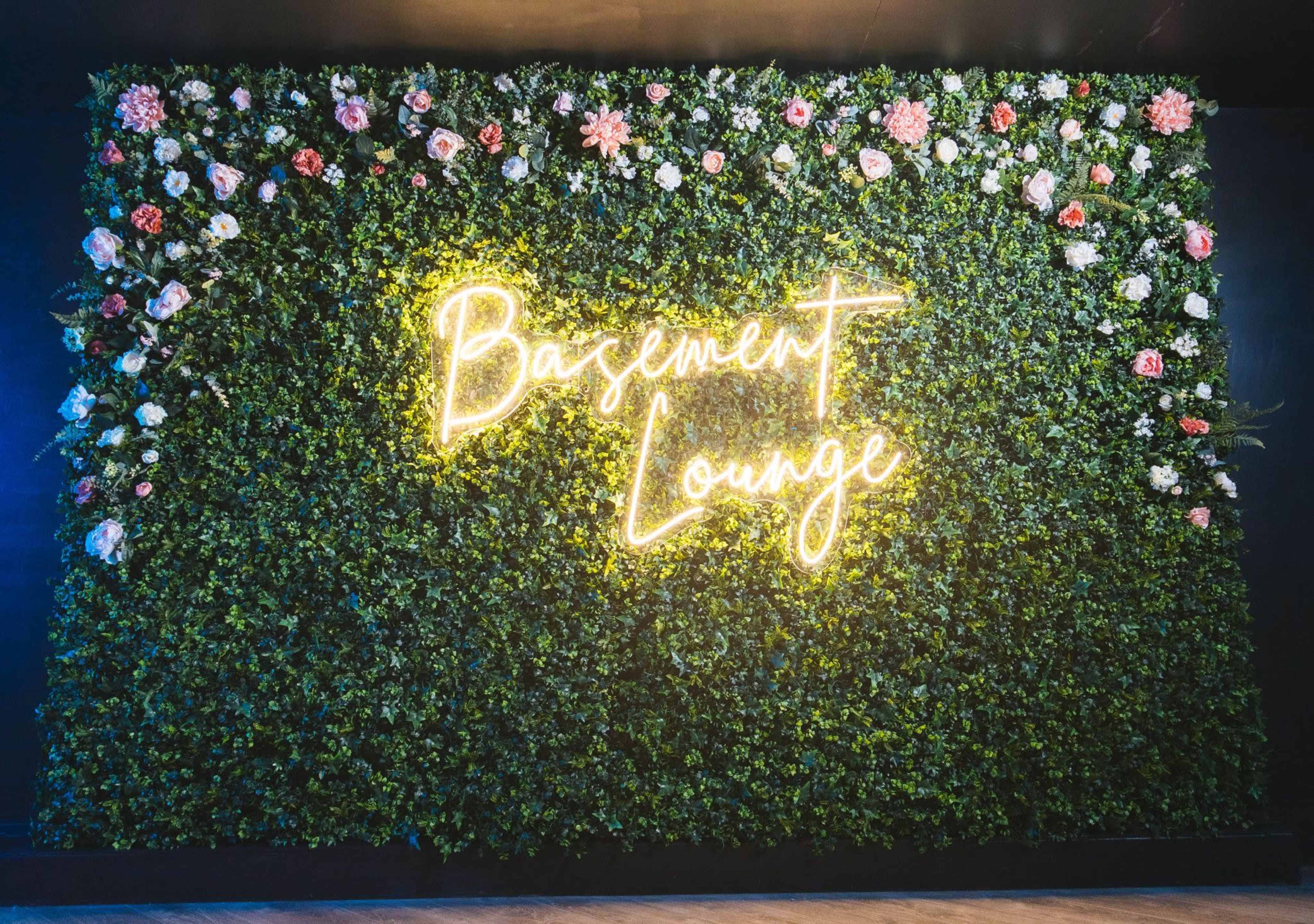 The image features a wall covered in artificial greenery with a neon sign that reads "Basement Lounge" illuminated in yellow.