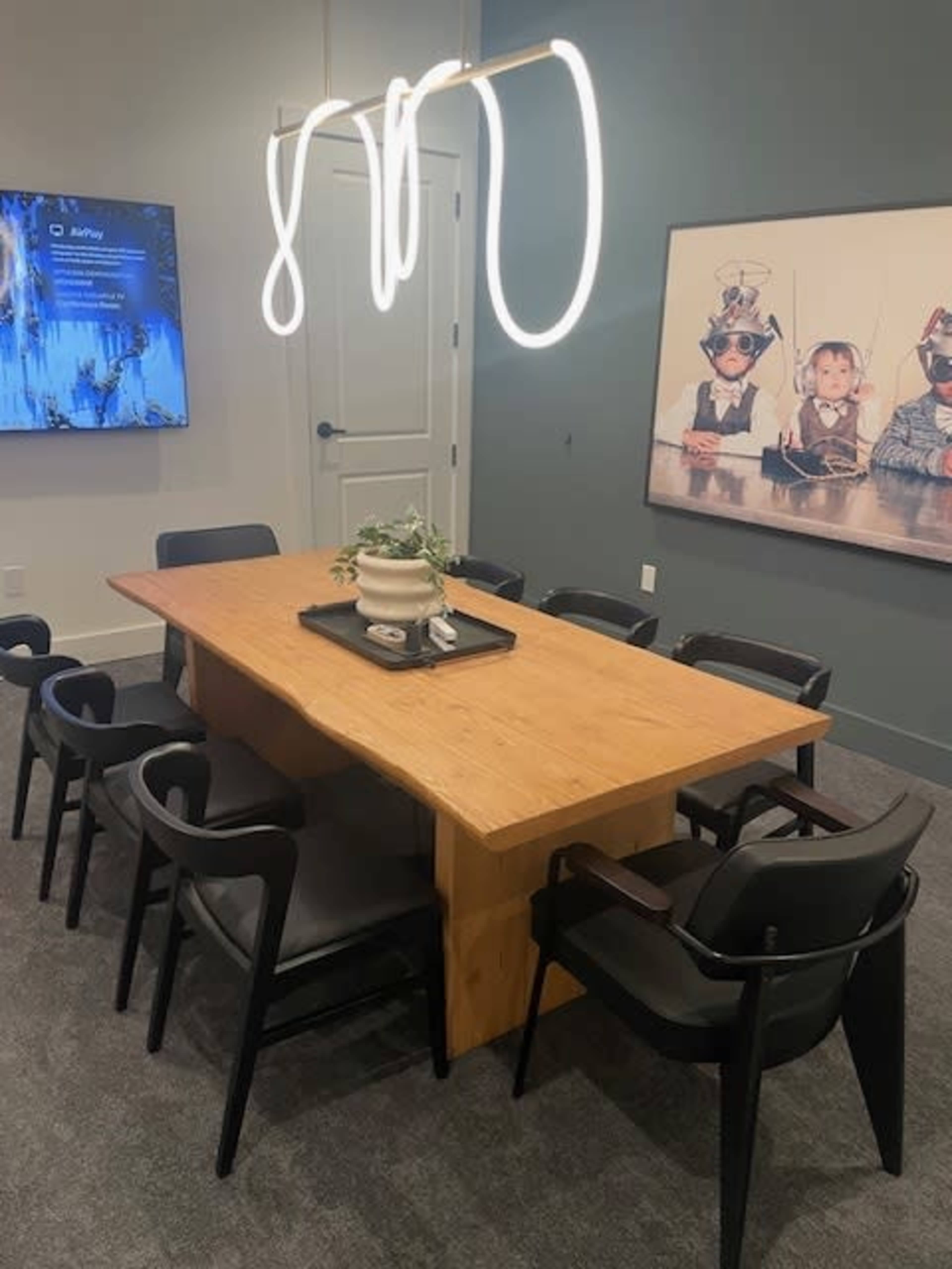 A modern conference room with a large wooden table surrounded by black chairs, a decorative plant on the table, and a wall featuring a framed photograph of children in playful costumes.