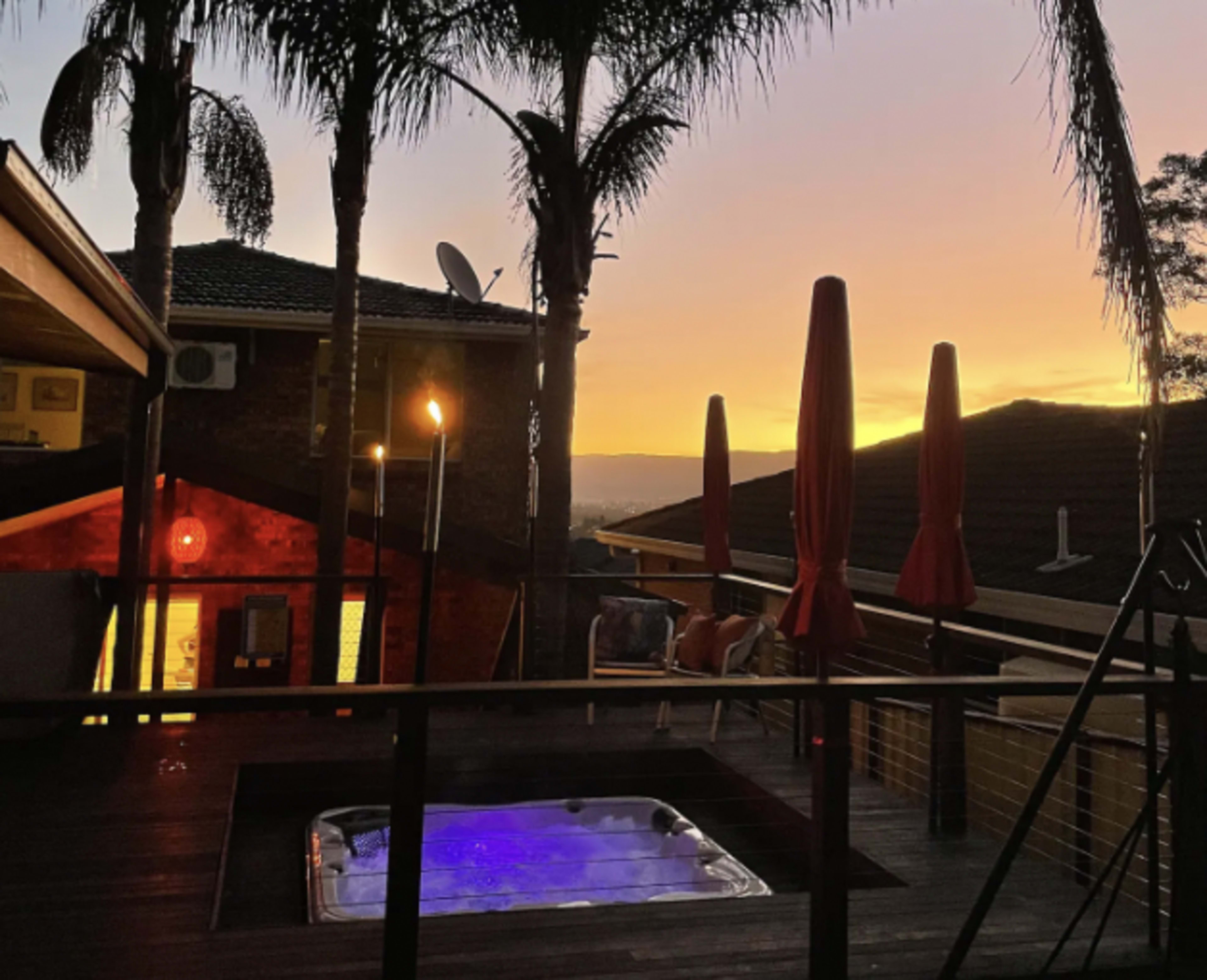 A hot tub on a deck surrounded by palm trees at sunset, with an adjacent building and red umbrellas nearby.